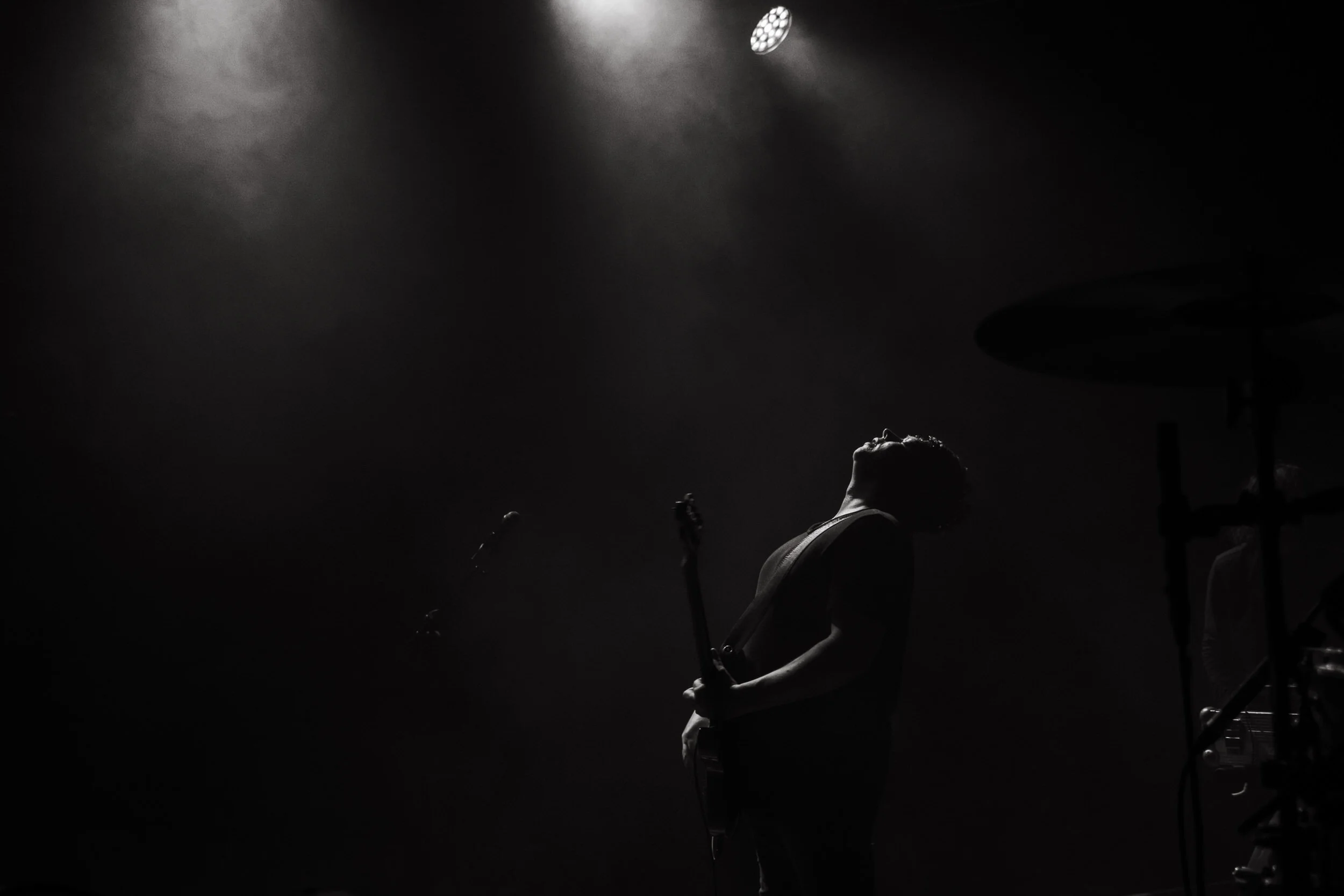 A musician performs on stage under a spotlight, head tilted back, with musical instruments and equipment visible around.