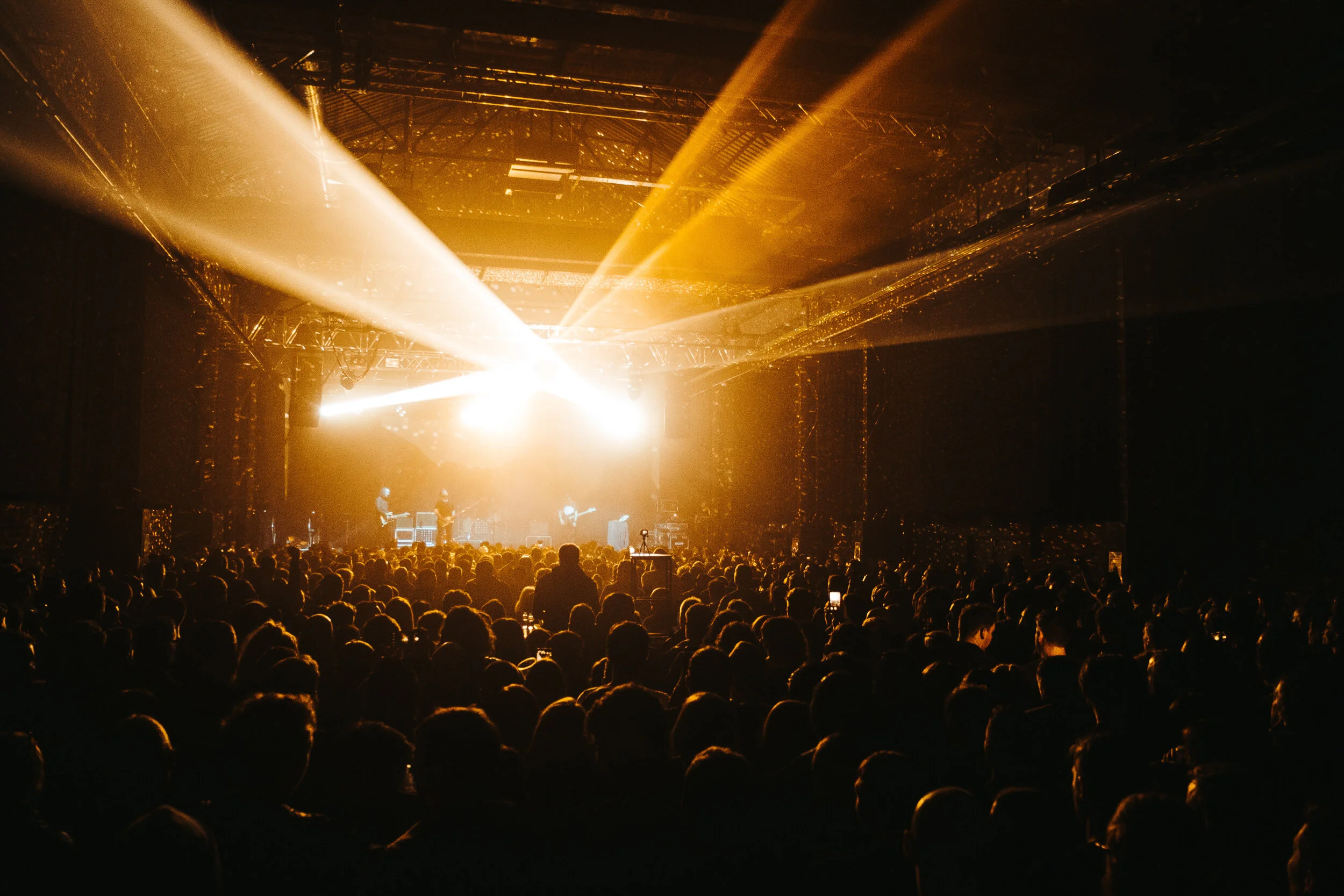 A concert with bright yellow and white stage lights shining over a large audience, with band members performing on stage.