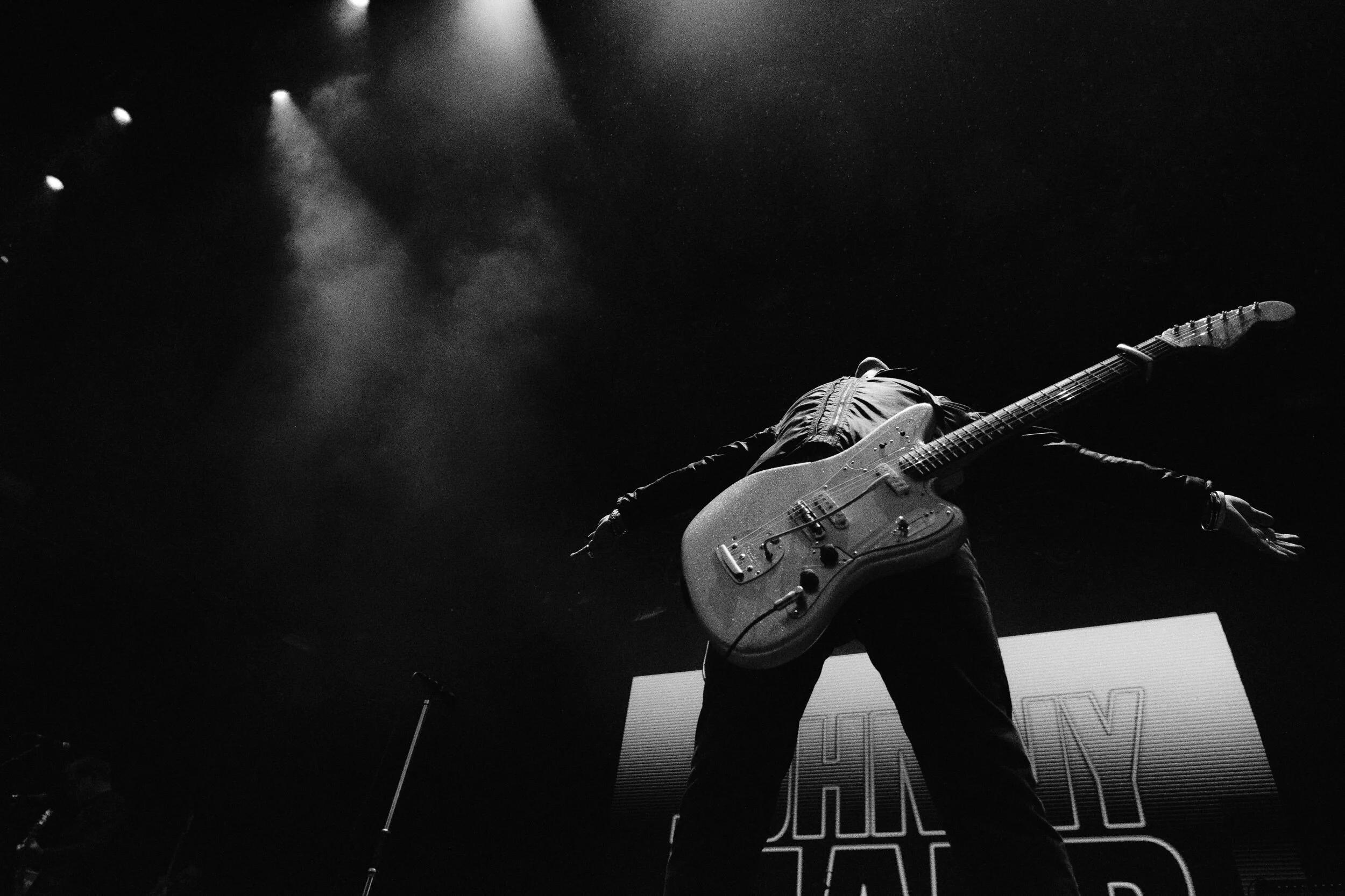 A musician on stage playing an electric guitar with a dark background and stage lighting.