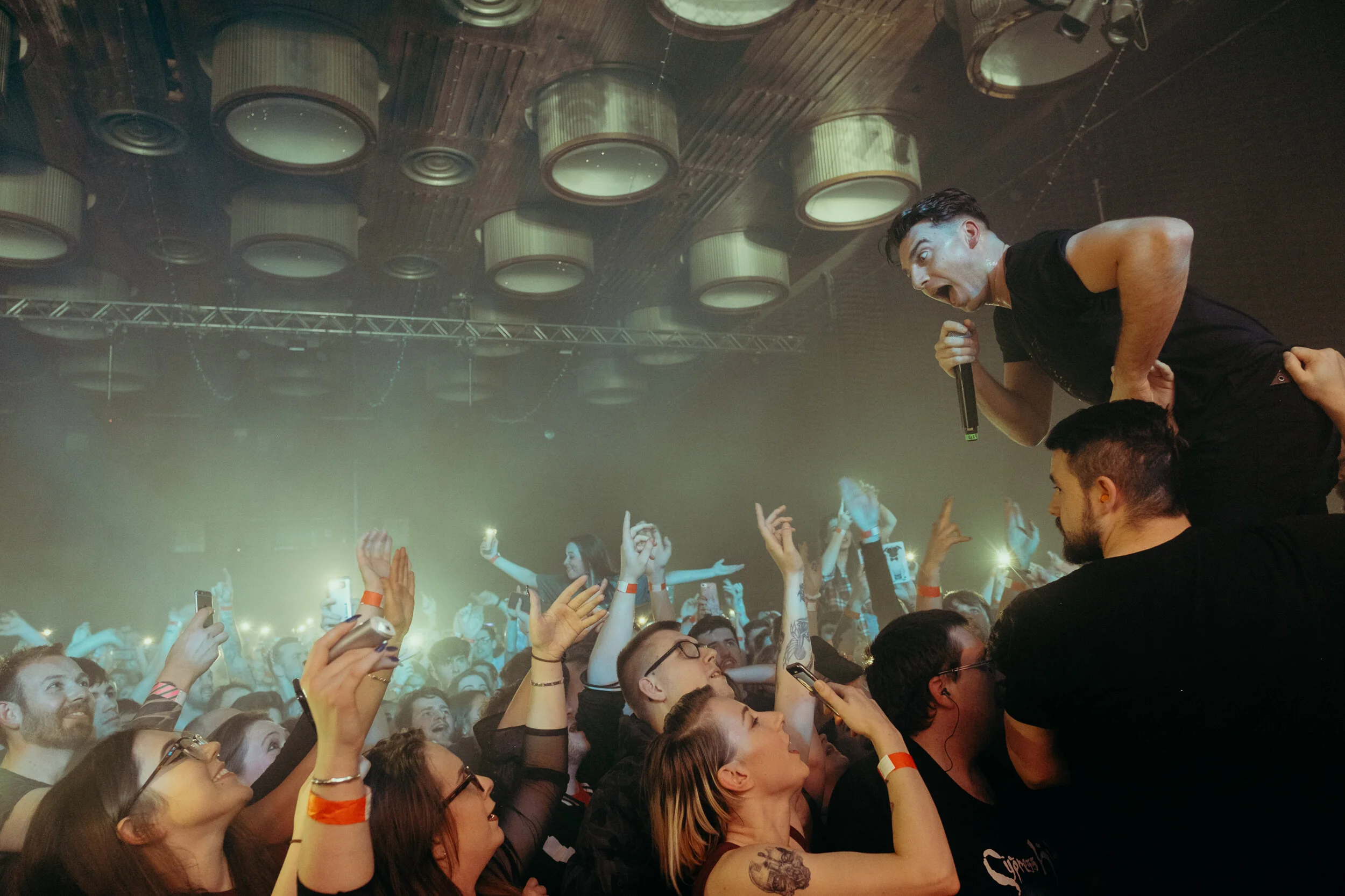 A concert scene with a singer holding a microphone, engaging with the crowd. The audience is excited, with many raising their hands and taking photos. The concert is indoors, with visible ceiling lights and equipment.
