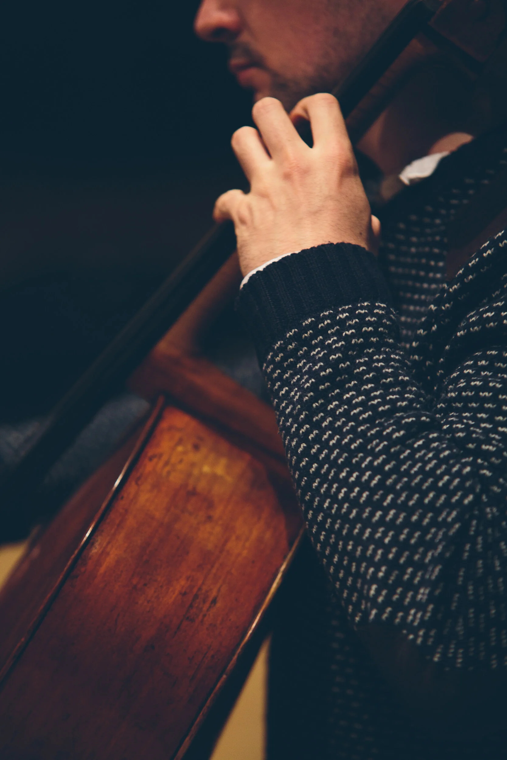 A person with a beard plays a string instrument, likely a cello, in a dimly lit setting, wearing a dark sweater with a white pattern.