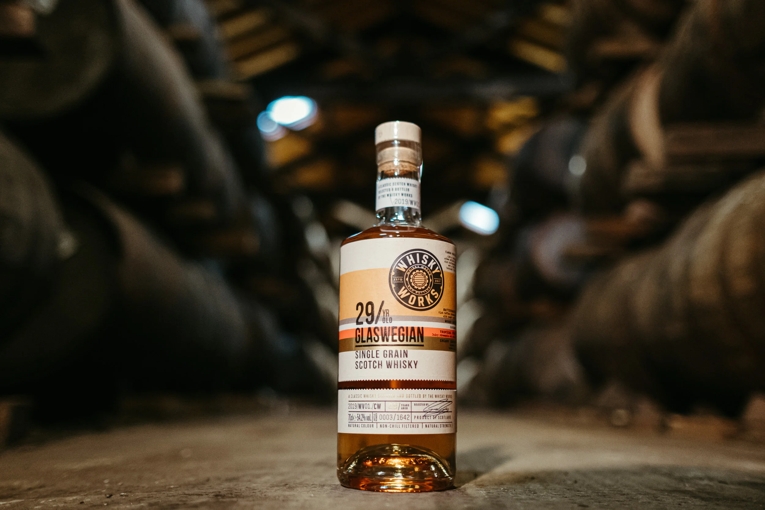 Bottle of Scotch whisky with a white label, placed on the floor in an underground wine cellar or storage area with shelves of wine bottles overhead.