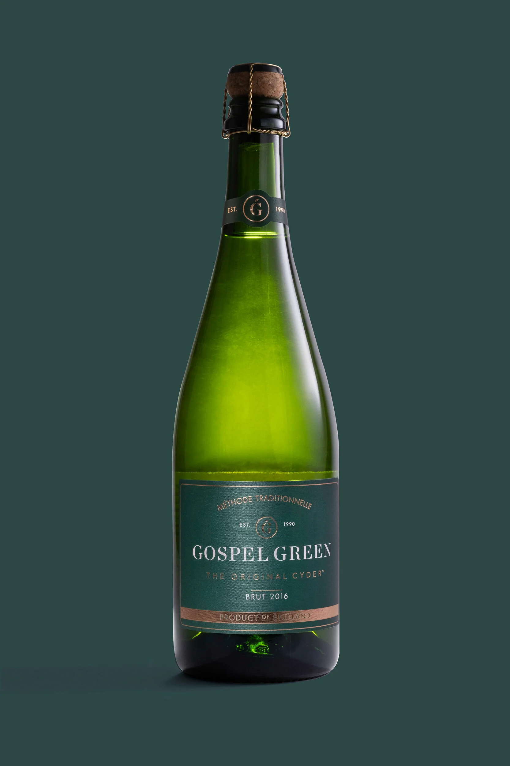 A green glass bottle of Gospel Green cider with a cork stopper secured by a wire cage, against a dark green background.