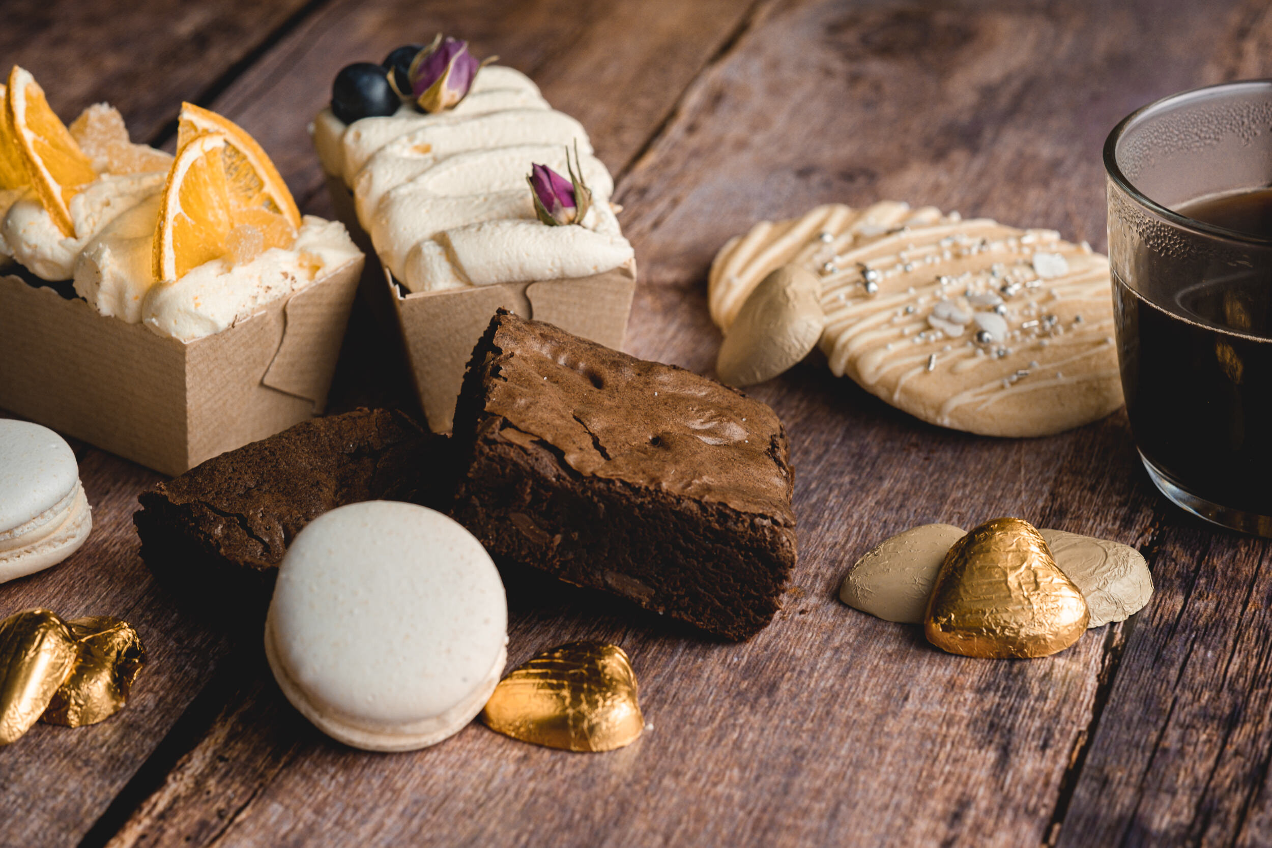 Assorted baked goods including cookies, brownies, and pastries, with a glass of coffee on a wooden surface, accented with gold foil-wrapped chocolates.