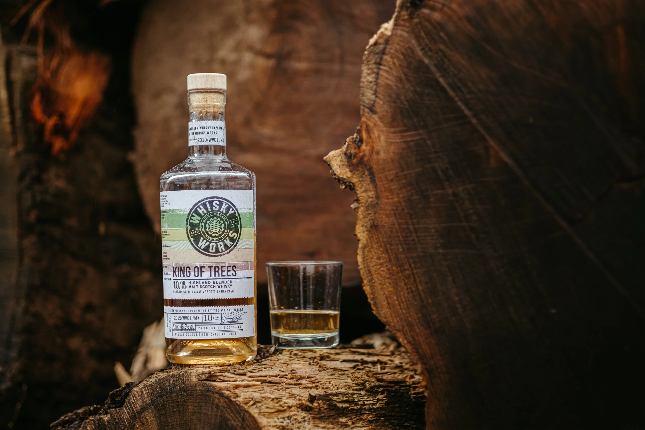 A bottle of whiskey, a small glass of whiskey, and a wood log background.