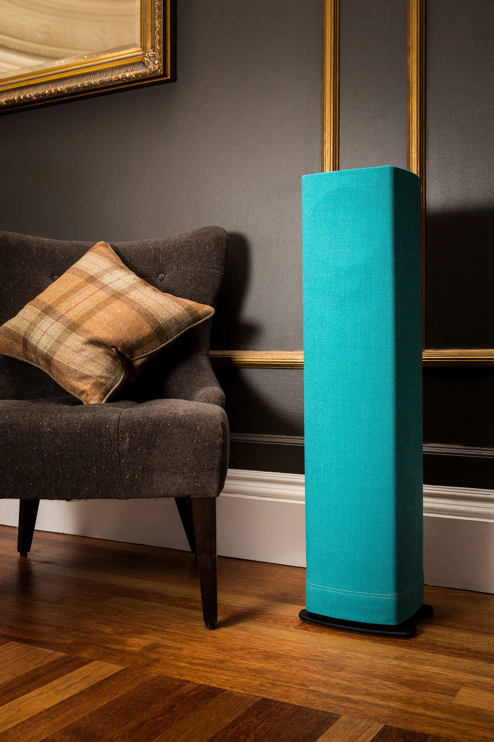 A living room scene with a brown upholstered armchair with a plaid cushion, a dark wall with a gold frame mirror, and a tall turquoise speaker or art piece on a black base on a wooden floor.