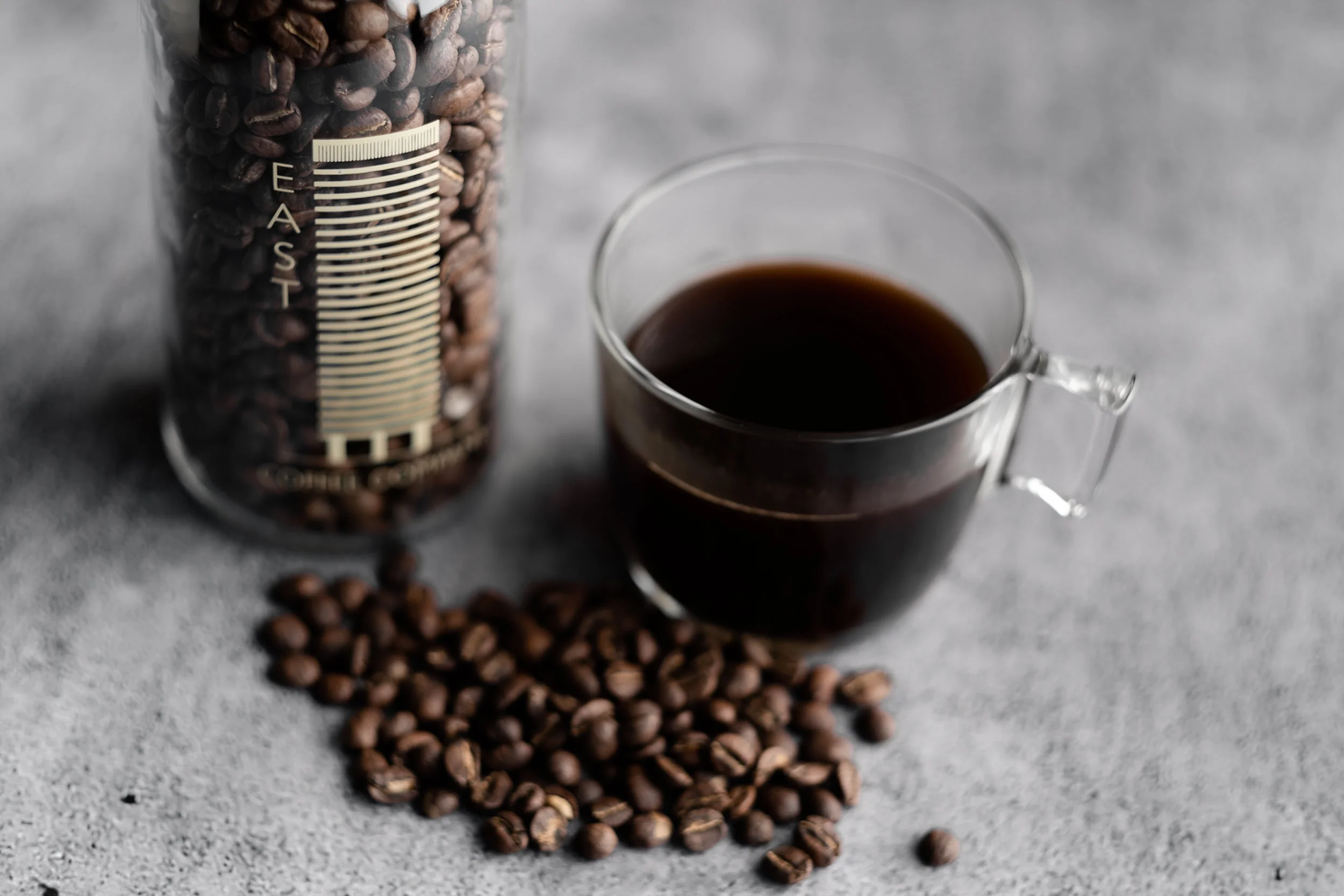 A clear glass jar filled with roasted coffee beans next to a small glass cup of black coffee on a gray surface, with some coffee beans scattered around.