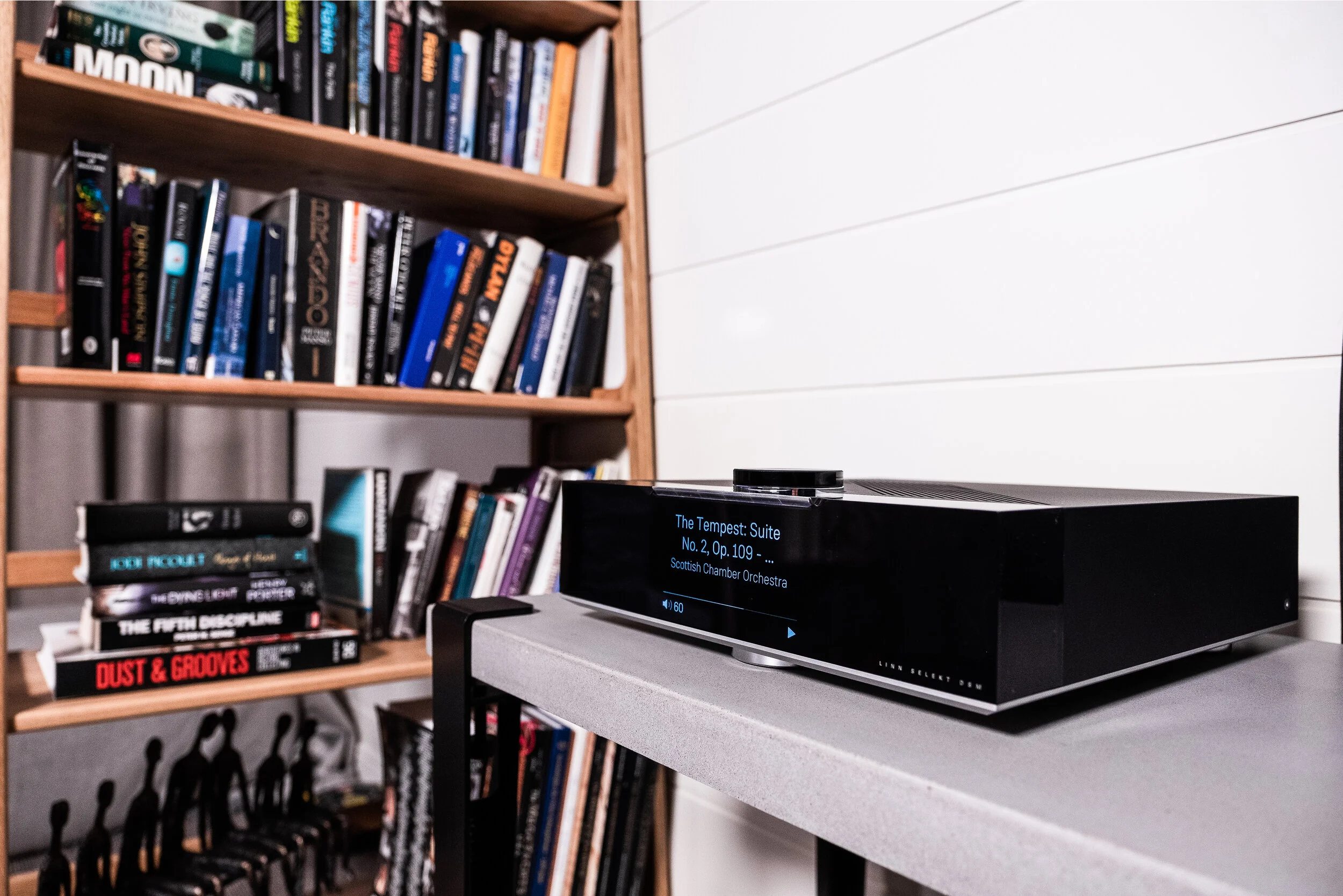 A music album cover with a black background and blue and white text displaying 'The Tempest: Suite No. 2, Op. 109 - Scottish Chamber Orchestra' on a black digital device, placed on a gray table in front of a wooden bookshelf filled with various books