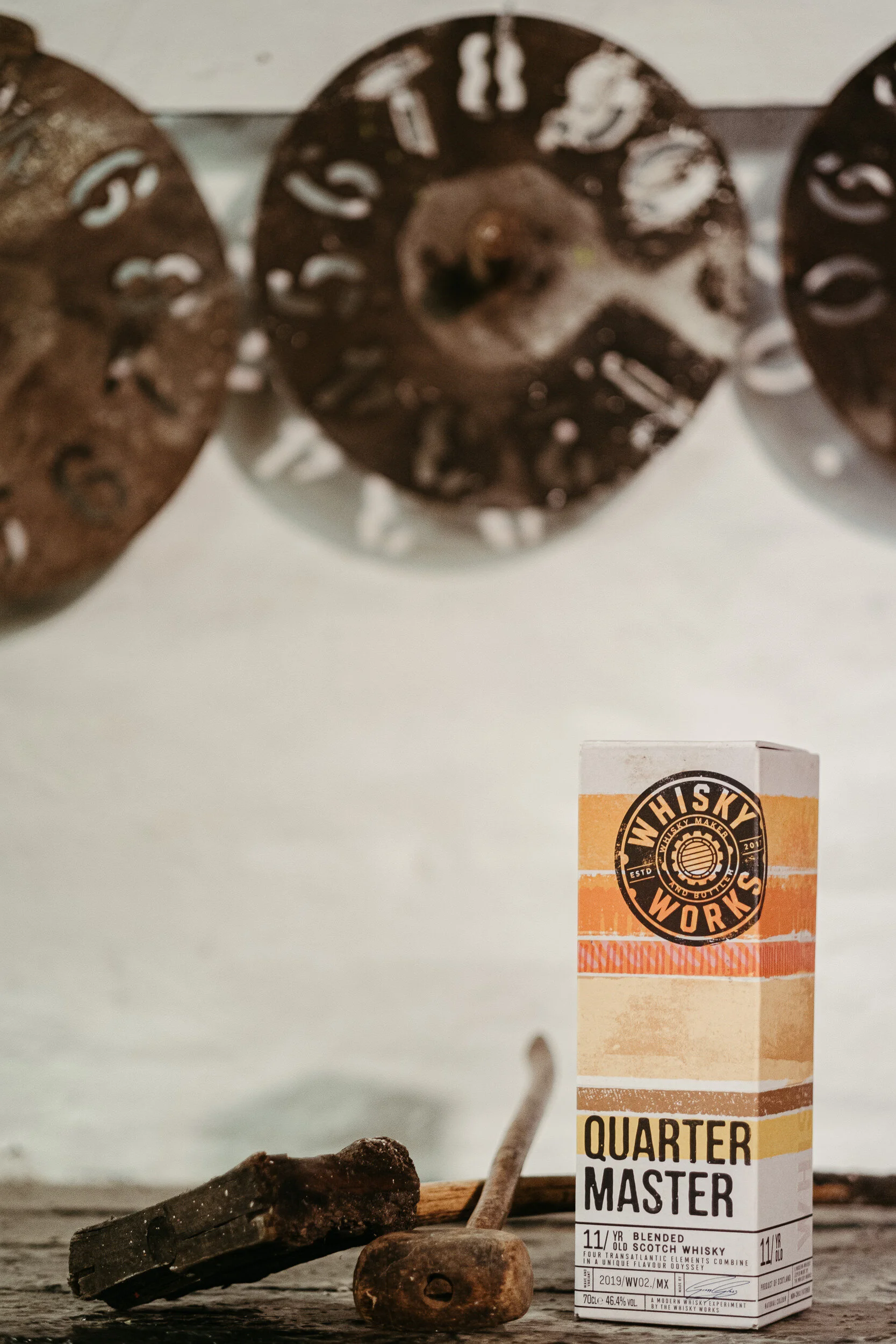 A box of Quarter Master blended old Scotch whisky on a rustic wooden surface with wood and metal objects nearby.