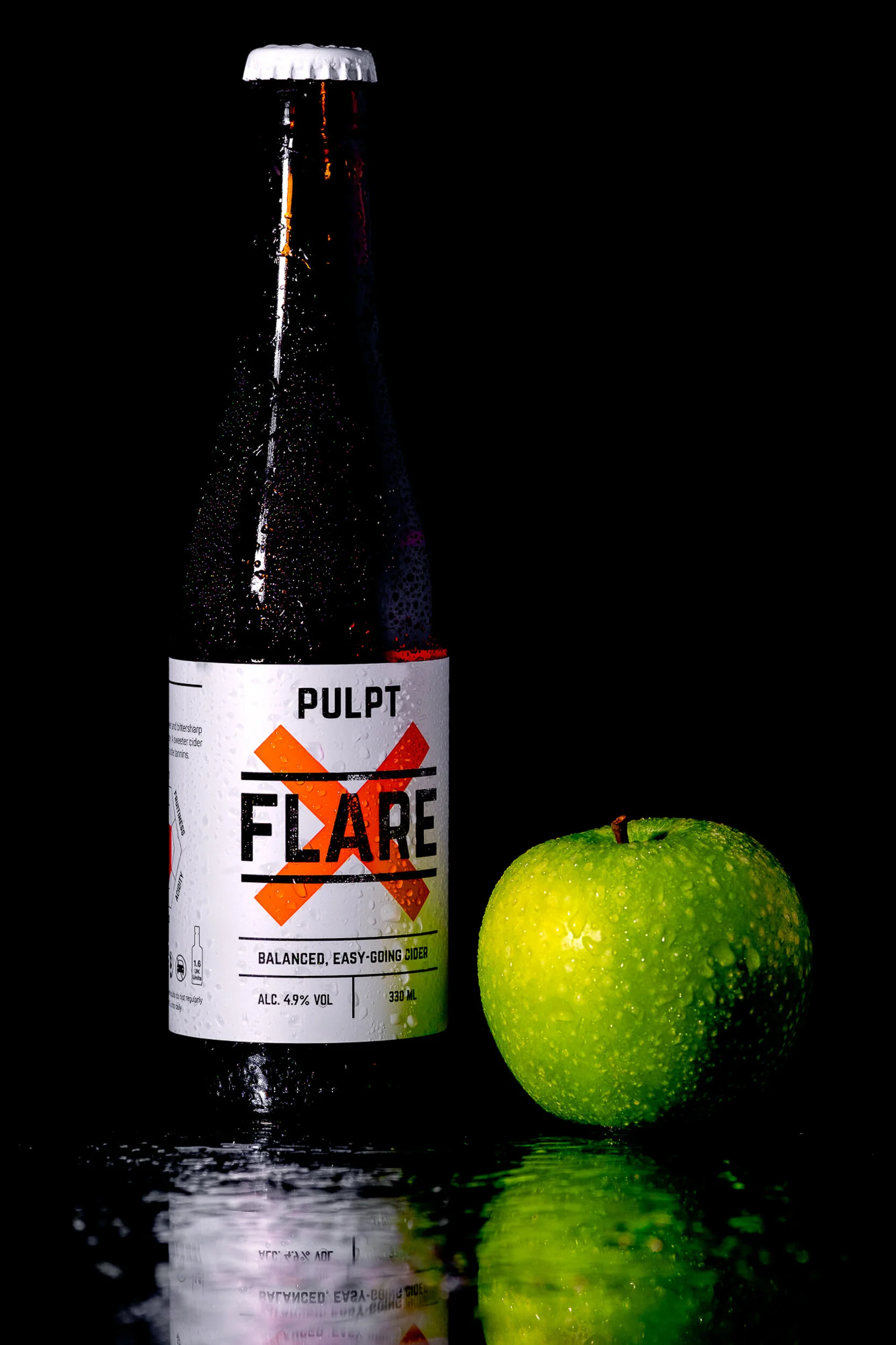 A green apple with water droplets next to a bottle of Pulp Fizz cider, against a black background with a reflective surface.
