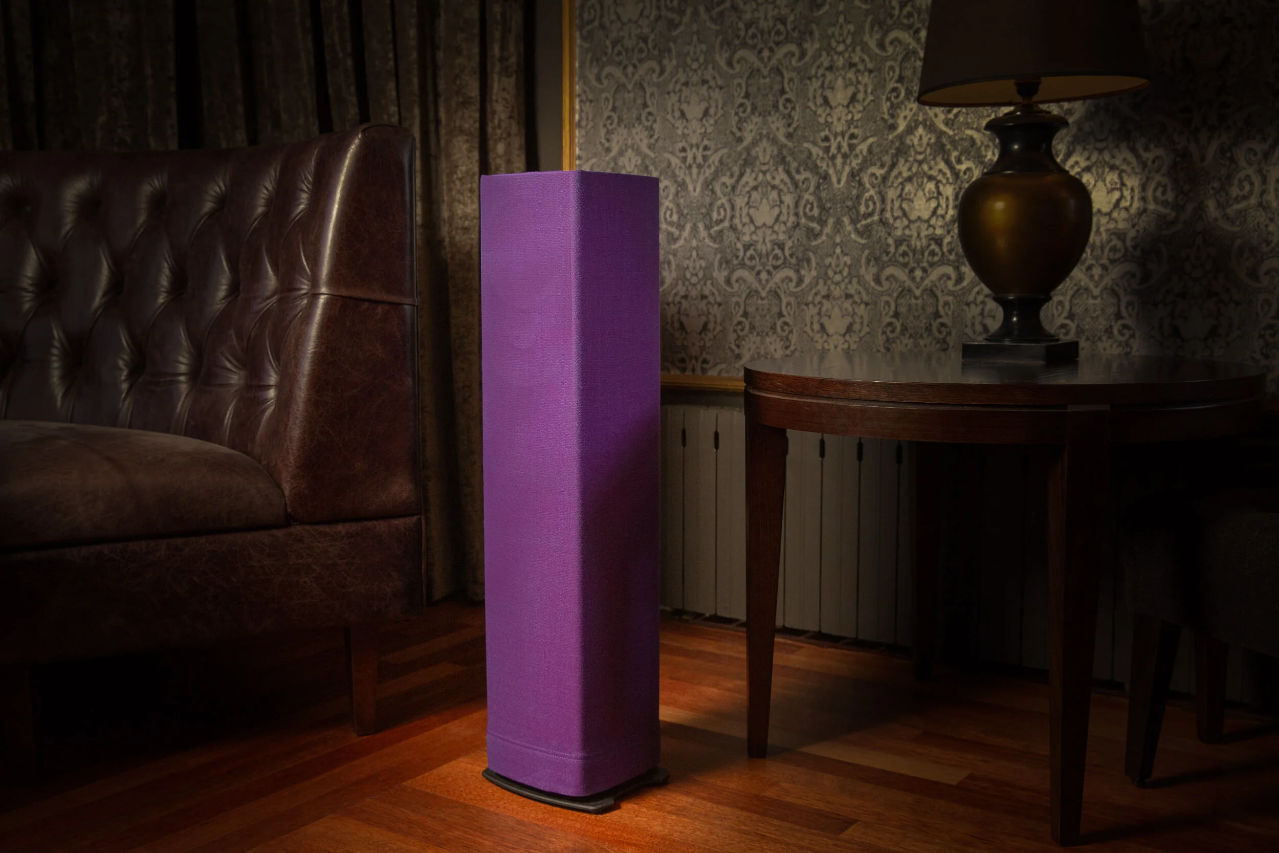 A tall purple speaker standing on a wooden floor next to a brown leather tufted armchair, with a lamp on a round wooden table in a room with patterned wallpaper and curtains.