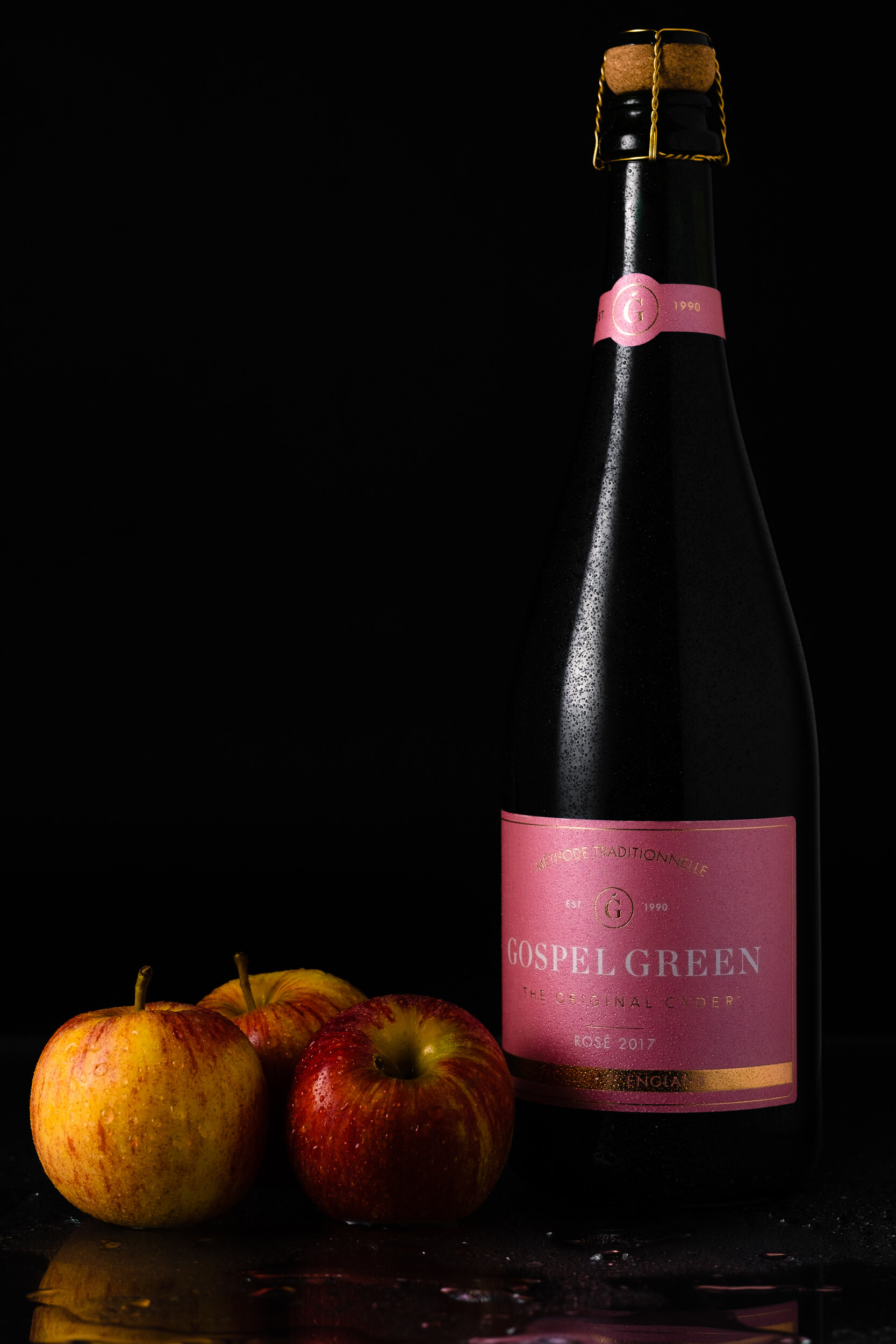 A black bottle with a pink label and a cork stopper, placed on a black surface with droplets of water, alongside four apples with red and yellow skin, with a dark background.