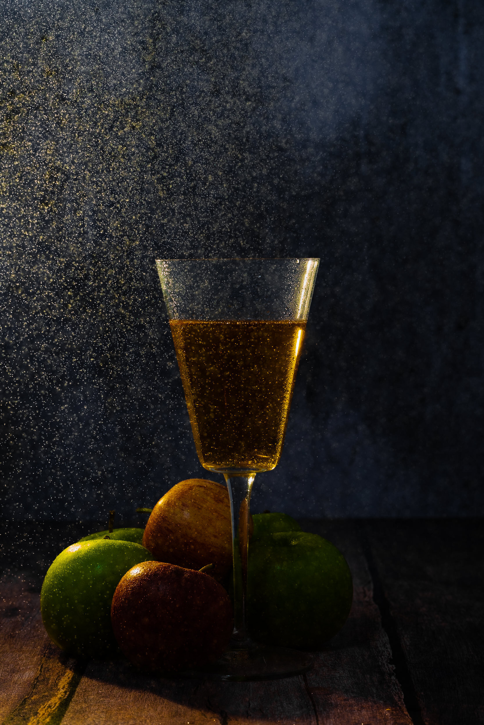 A champagne flute filled with sparkling champagne, with remnants of bubbles airborne for a celebratory atmosphere. Several apples, both green and red, are arranged at the base of the glass on a rustic wooden surface, against a dark background.