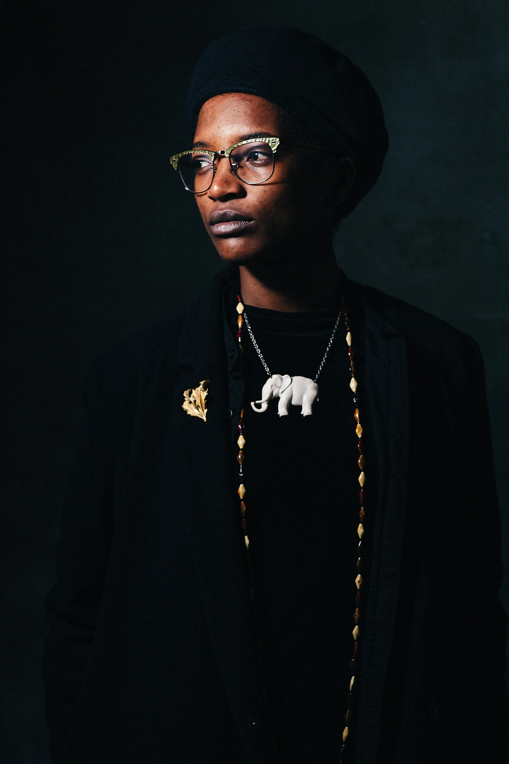 A young person with short hair, wearing glasses, a black beret, and a black shirt. They have multiple necklaces, including one with a white elephant pendant and another with beads, and a gold leaf accessory on their shirt. The background is dark, hig