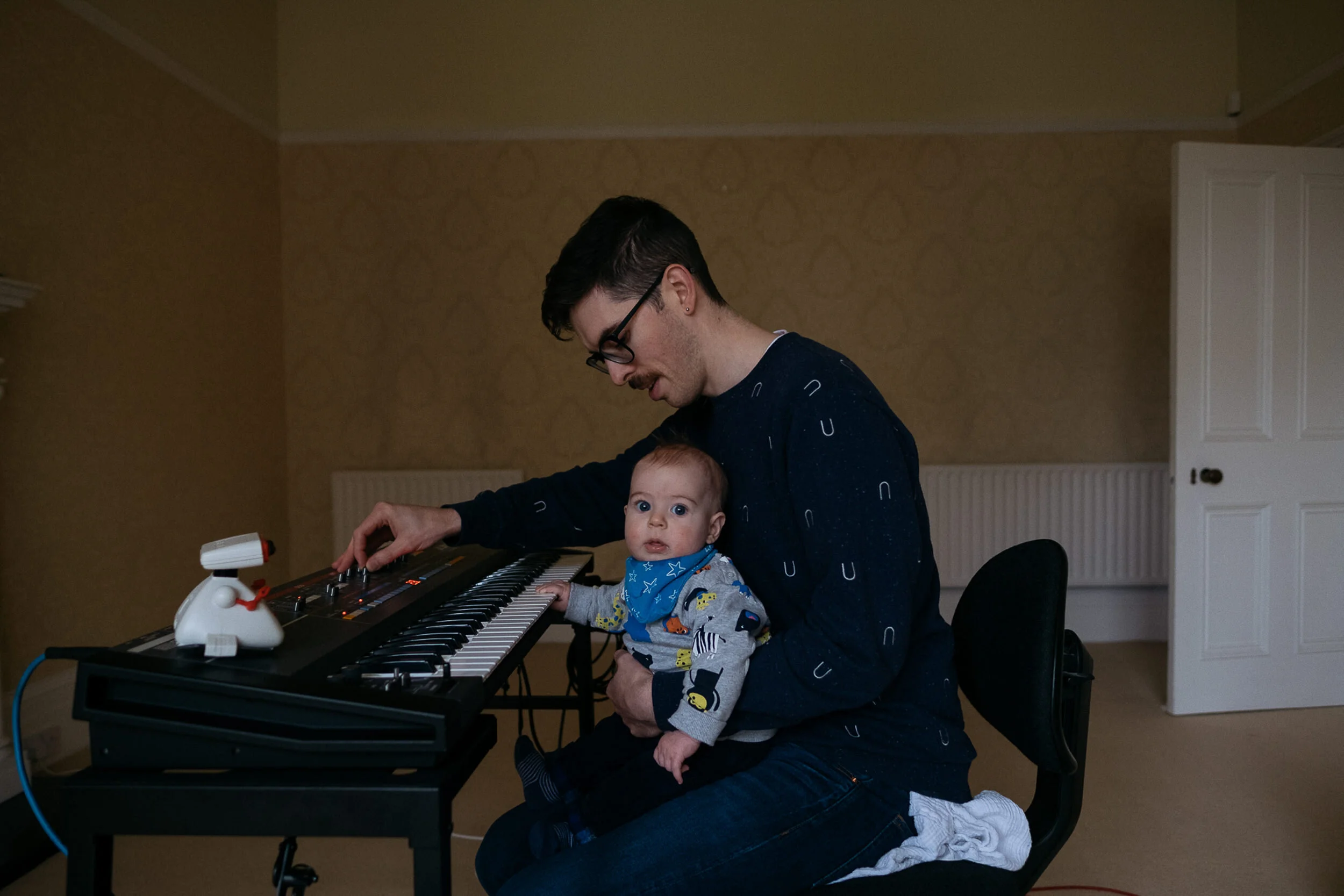 A man with glasses and dark hair playing an electronic keyboard with a baby sitting on his lap, looking at the camera, in a room with beige walls and a white door.