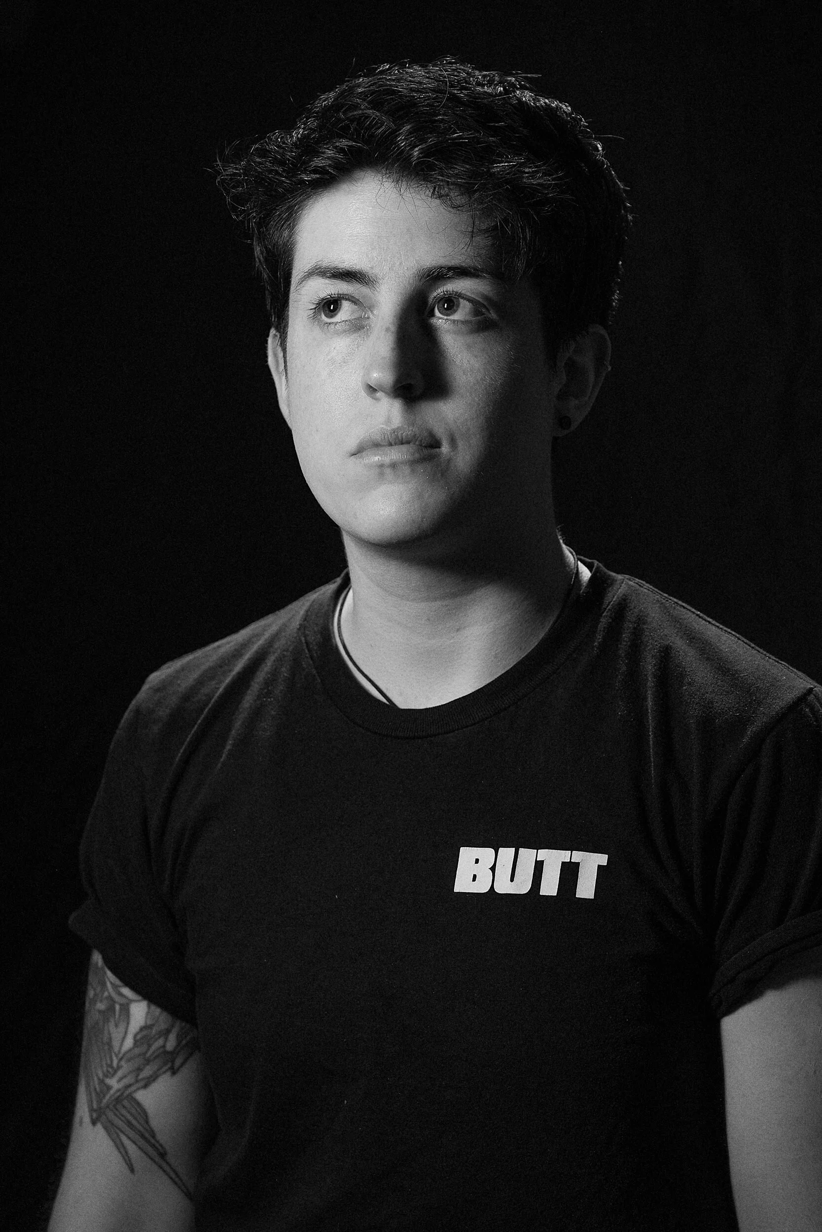 Black and white photo of a young man with short, dark, curly hair, wearing a black t-shirt with the word 'BUTT' printed on it, looking to the side, with partial arm tattoo visible.