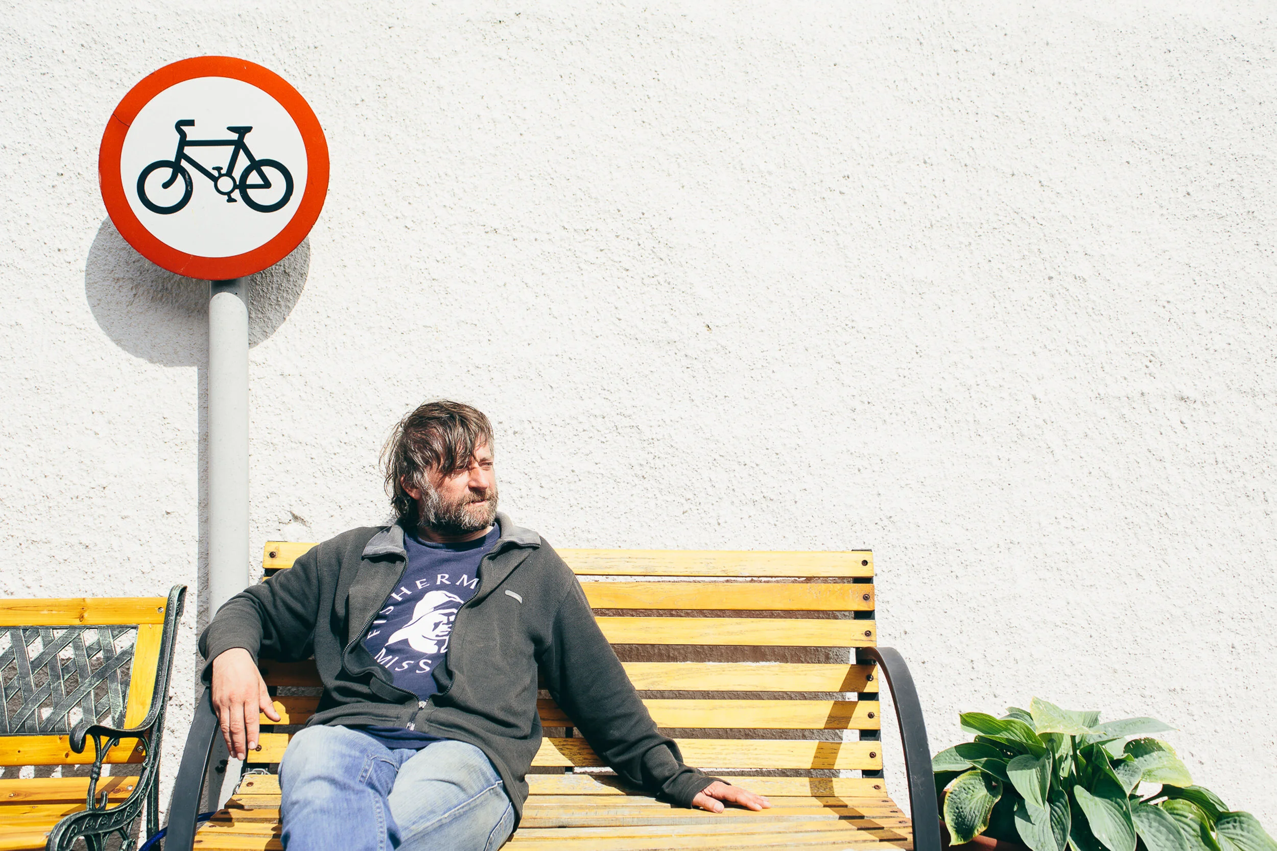 A man with brown hair, beard, and wearing a gray jacket and a T-shirt, sitting on a yellow bench with black armrests, outdoors against a plain white textured wall. A bicycle sign with a red circle is on the wall behind him, and there are green plants
