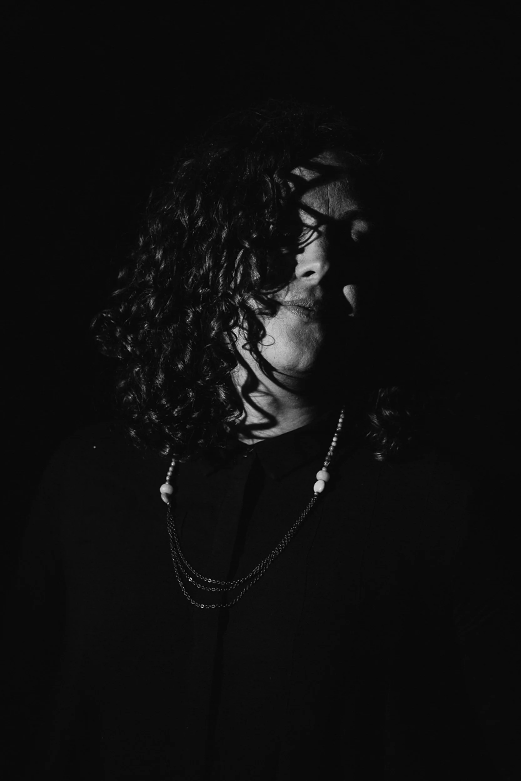 A black-and-white portrait of a woman with curly hair, wearing a dark shirt, pearl necklace, and layered chains, with her head lowered and eyes closed against a dark background.