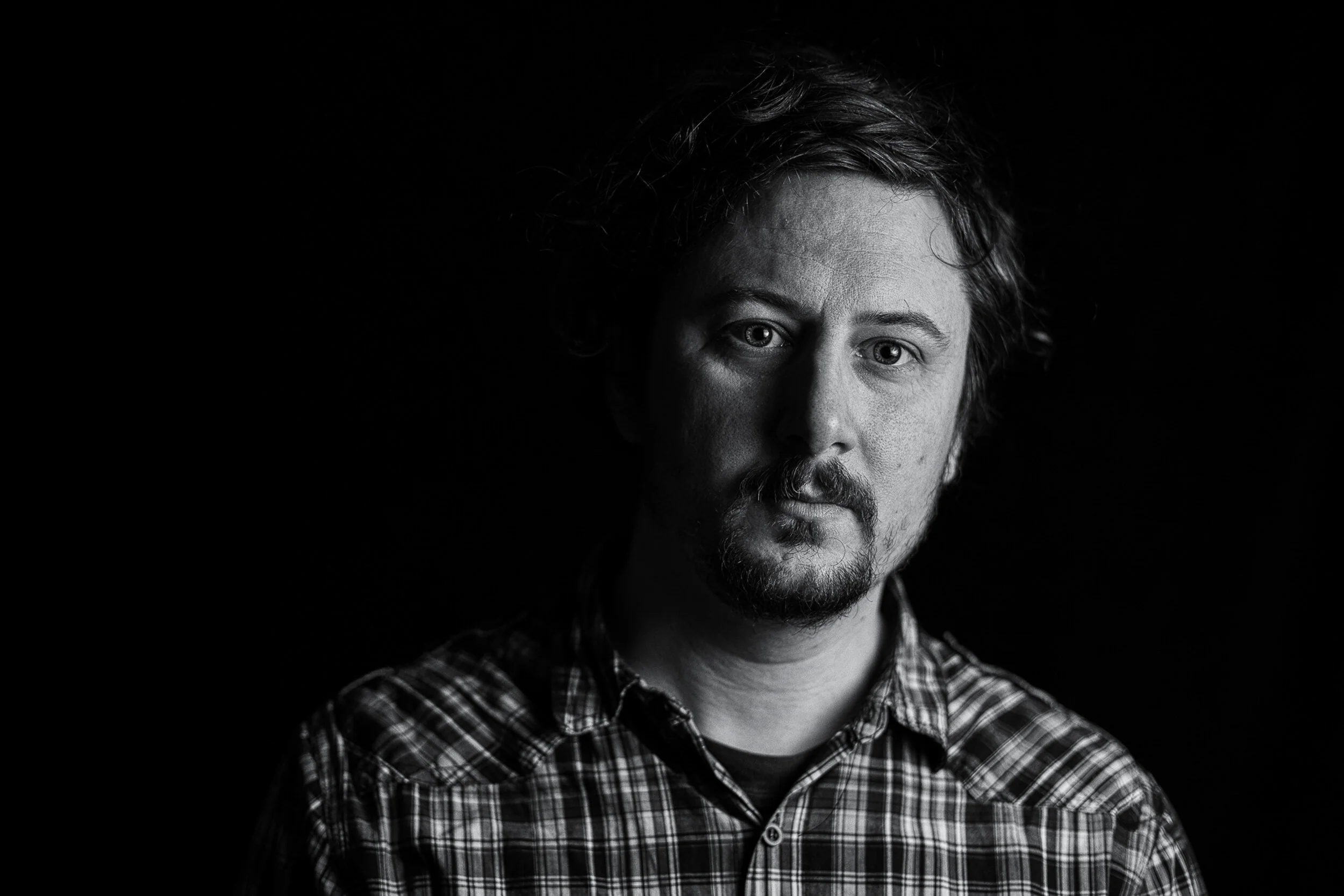 Black and white portrait of a man with curly hair and a beard, wearing a plaid shirt, against a dark background.