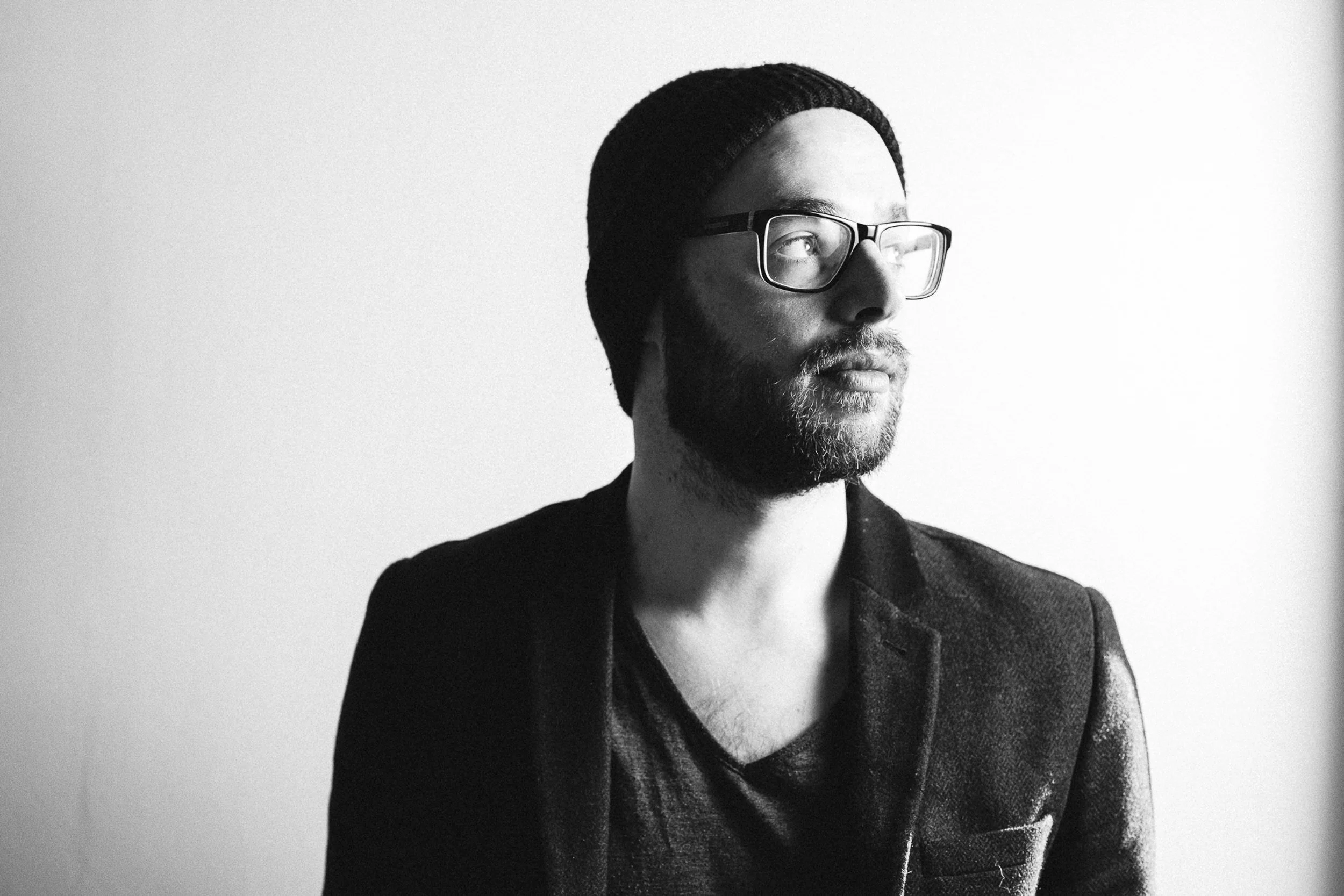 Black and white photo of a man with glasses, beard, mustache, wearing a beanie and a blazer, looking to the right.