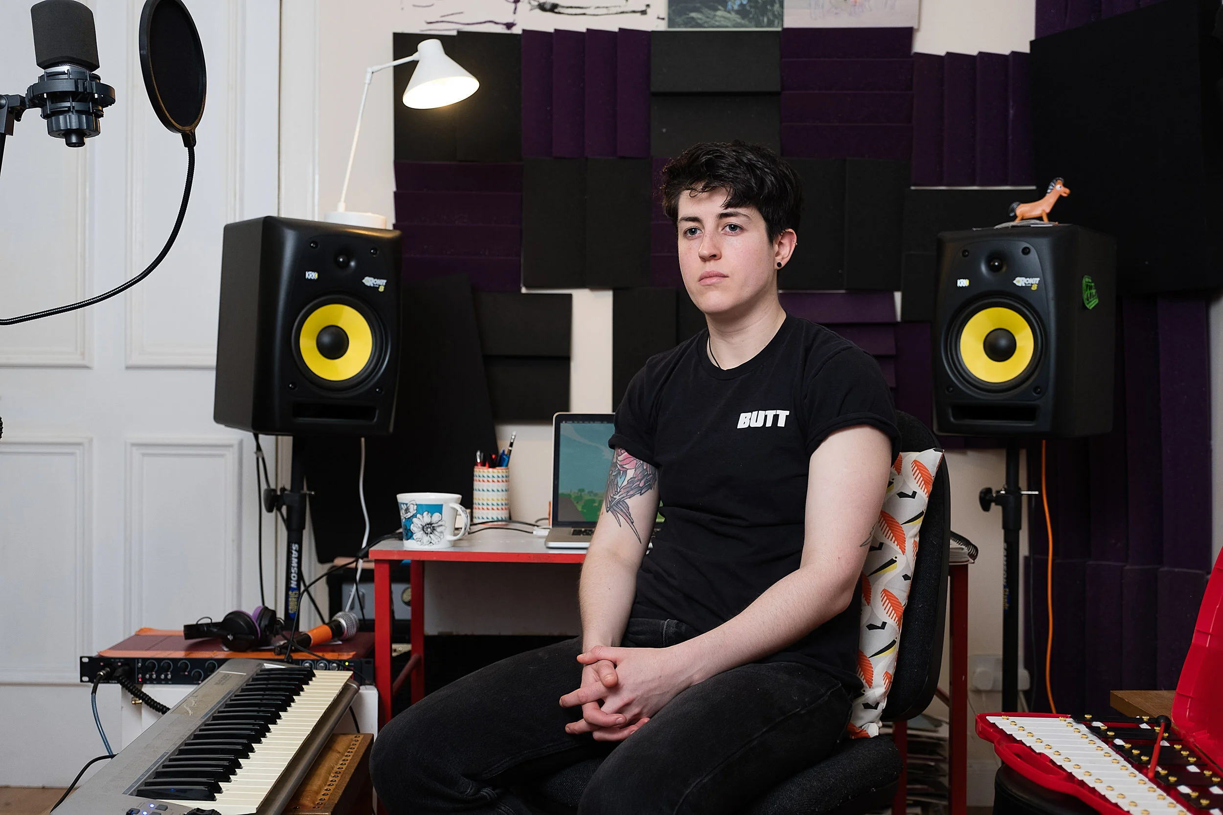A young person sitting in a music studio surrounded by electronic and acoustic musical equipment. There are two yellow speaker monitors, a keyboard, a microphone, a laptop, and other musical accessories. The room has black and purple soundproofing pa