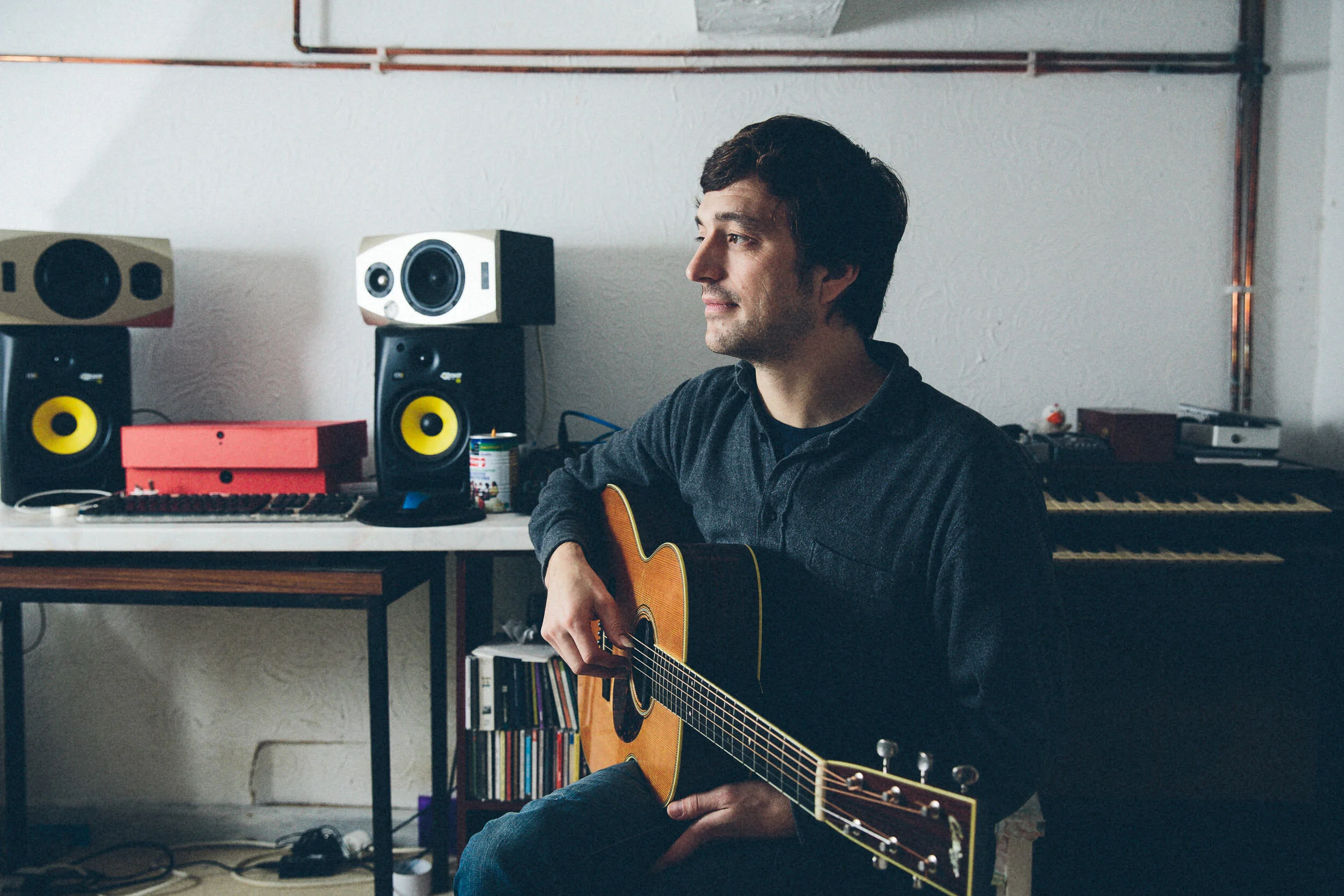 A man with dark hair playing an acoustic guitar in a music studio. The studio has two large studio monitors with yellow cones, a keyboard on a stand, bookshelves filled with CDs or DVDs, and a computer mouse on the desk.