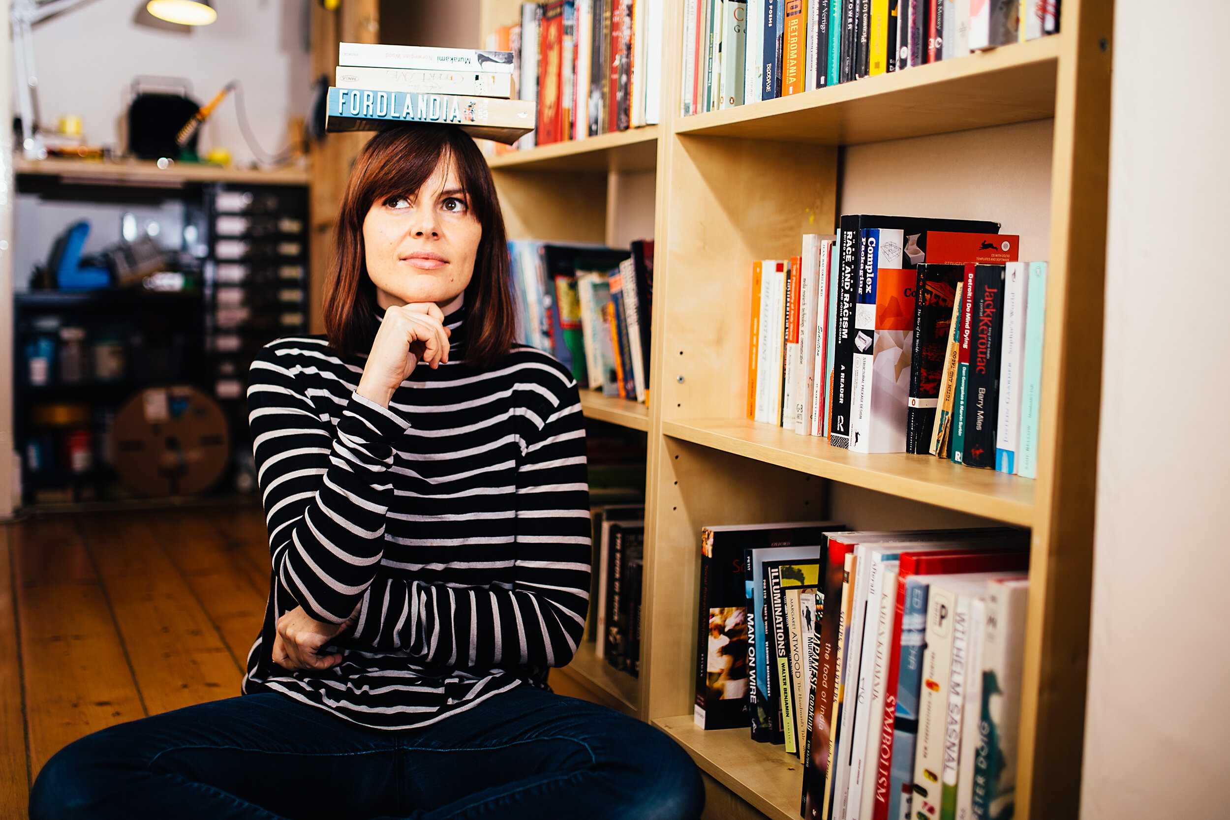 A woman with shoulder-length brown hair and wearing a black and white striped shirt is sitting on a round, dark blue cushion in a library or bookstore, balancing a row of books on her head while surrounded by shelves filled with colorful books.