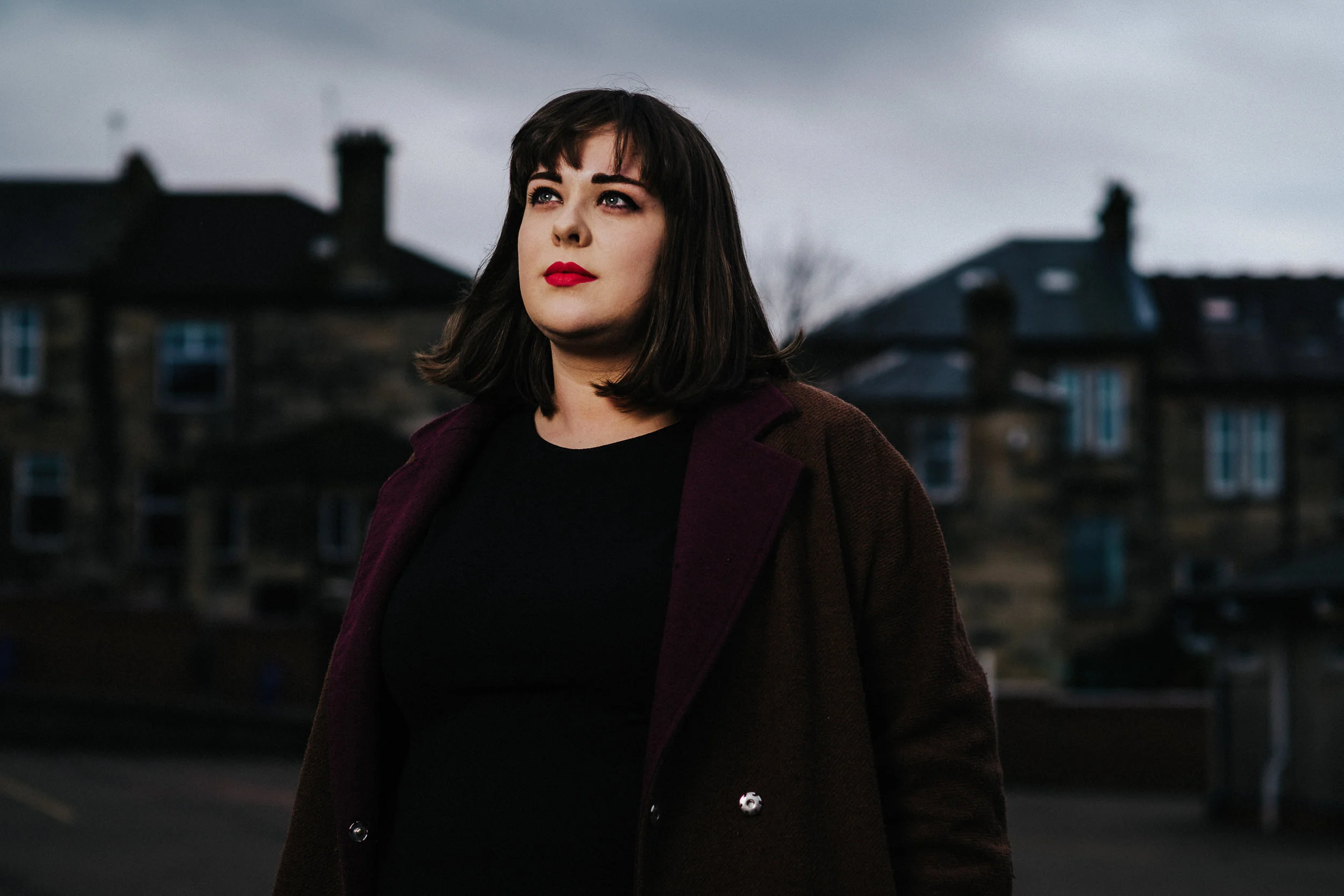 A woman with dark brown hair and red lipstick looking into the distance outdoors during overcast weather, with blurred buildings in the background.
