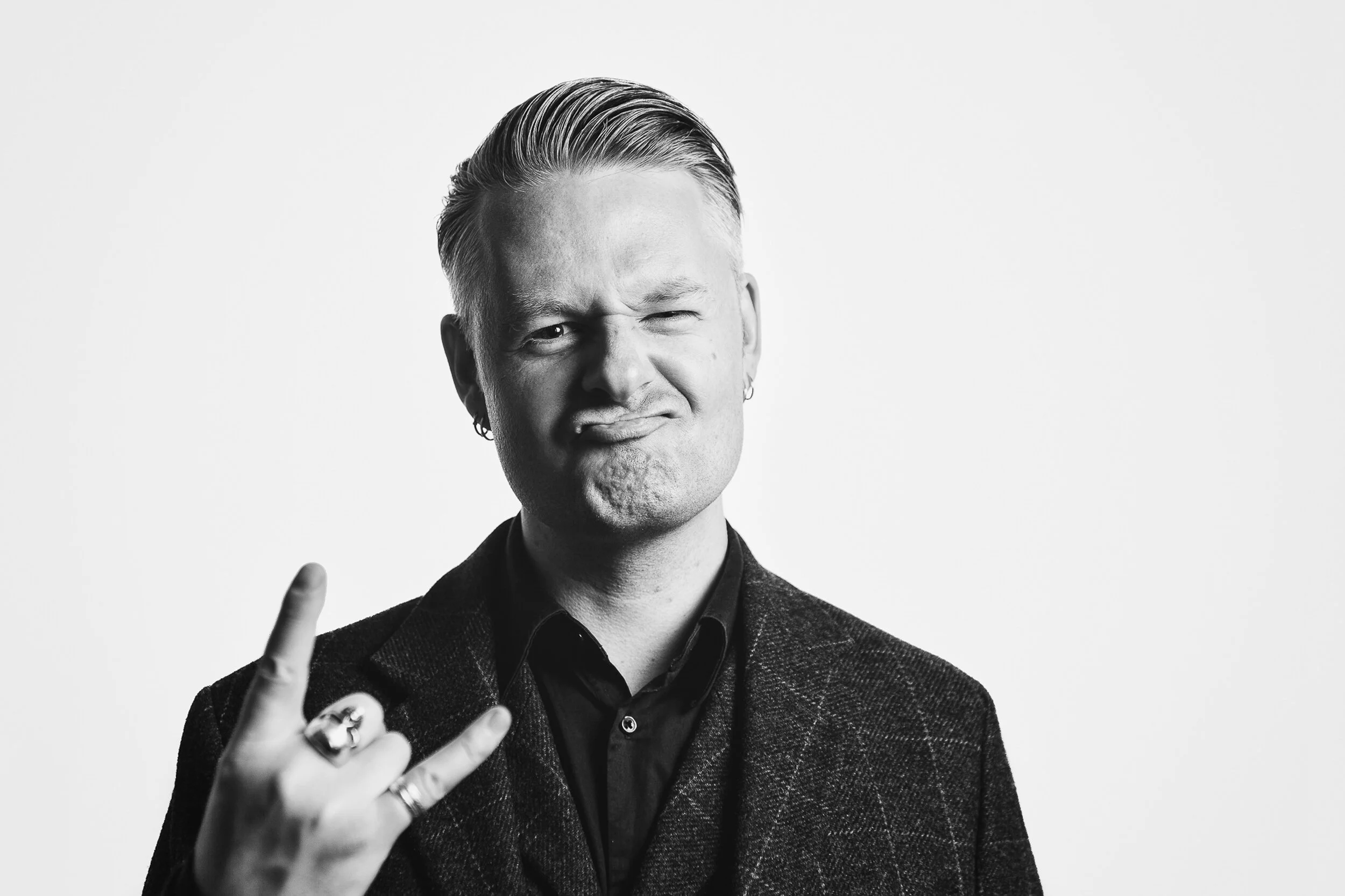 Black and white photo of a man with slicked-back hair making a face, winking with one eye and puckering his lips, while holding up his hand with fingers in a loose gesture.