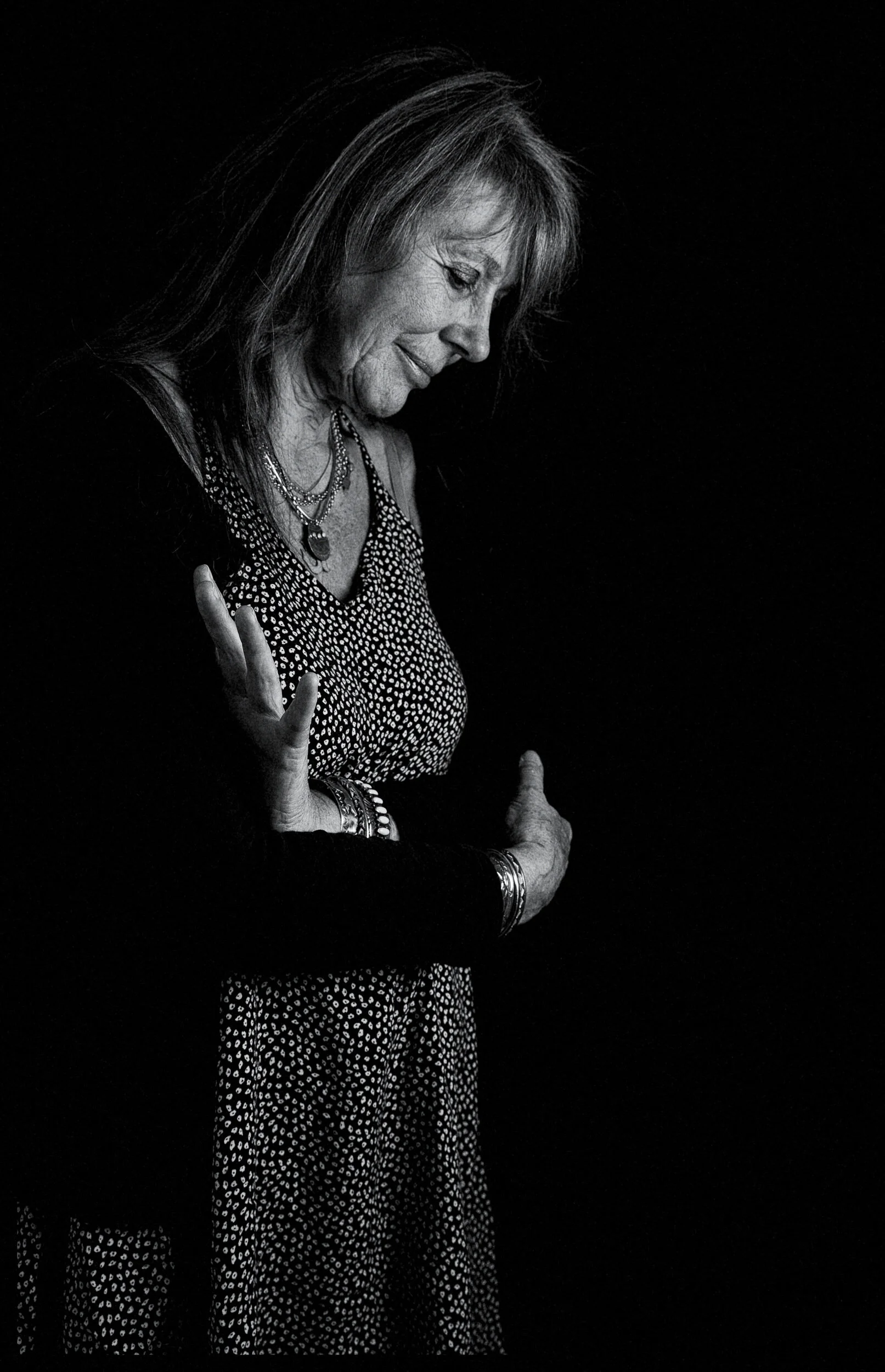 Black and white photo of an elderly woman with long hair, wearing patterned clothing and jewelry, making a gentle gesture with her hands, side profile, dark background.