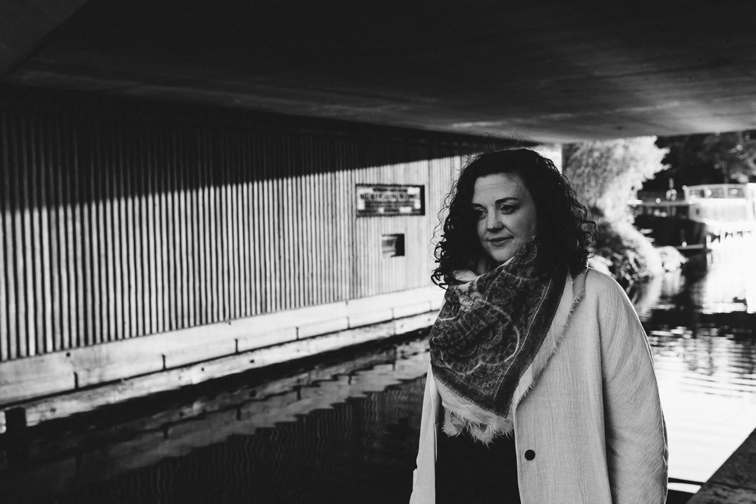 A woman with curly hair standing outdoors next to a body of water, wearing a coat and a scarf, with a bridge and trees in the background.