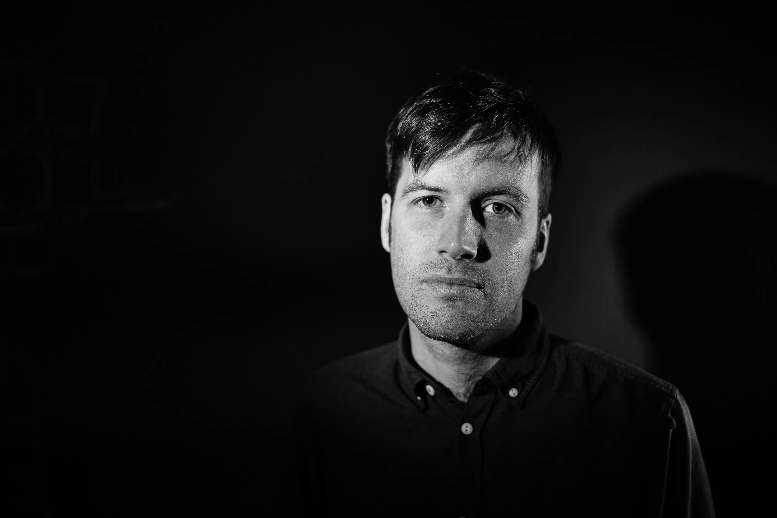 Black and white portrait of a man with short dark hair, wearing a dark collared shirt, looking at the camera against a dark background.