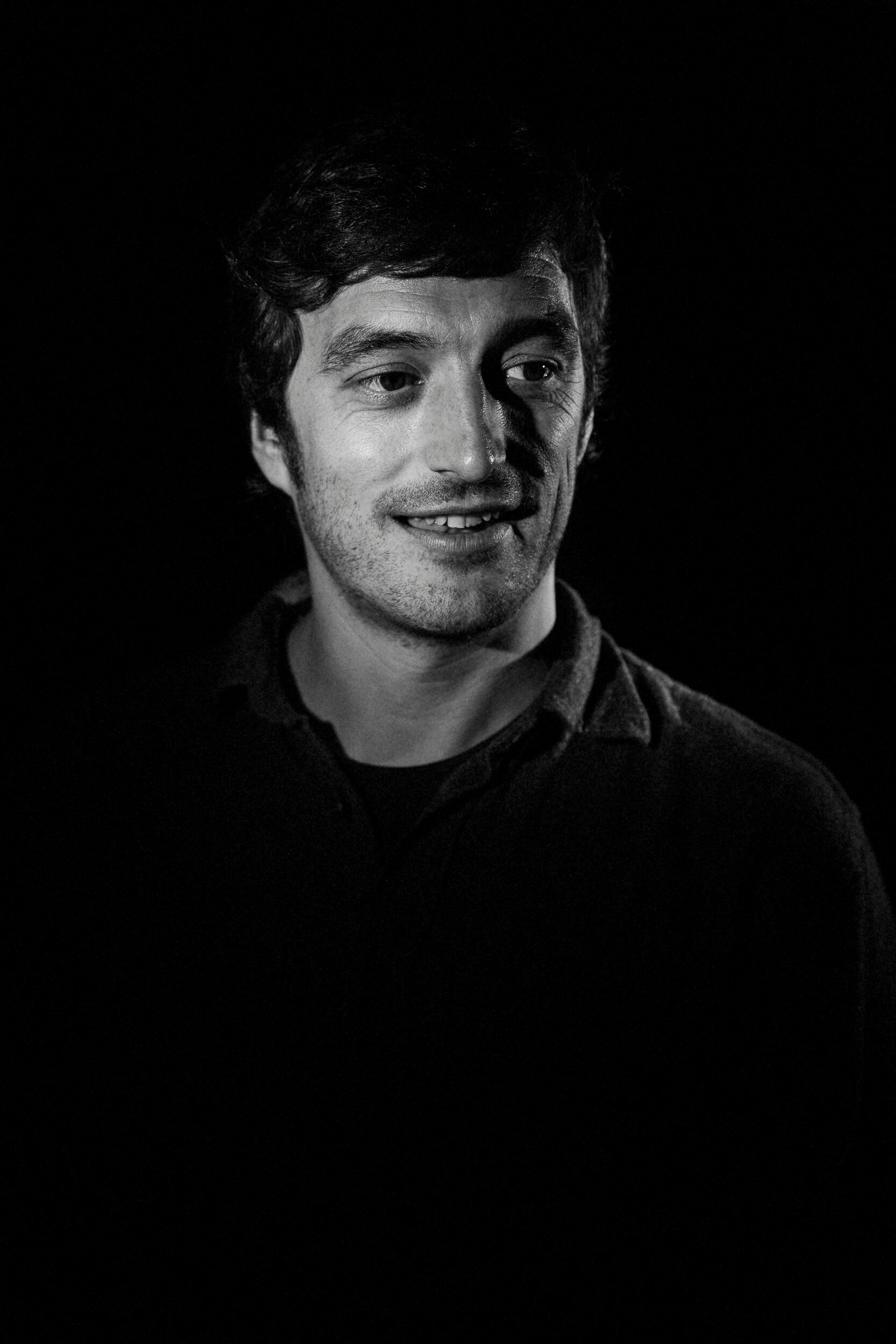 Black and white portrait of a man with dark hair, slight beard, smiling with mouth slightly open, wearing a dark shirt, facing slightly to the right, against a dark background.