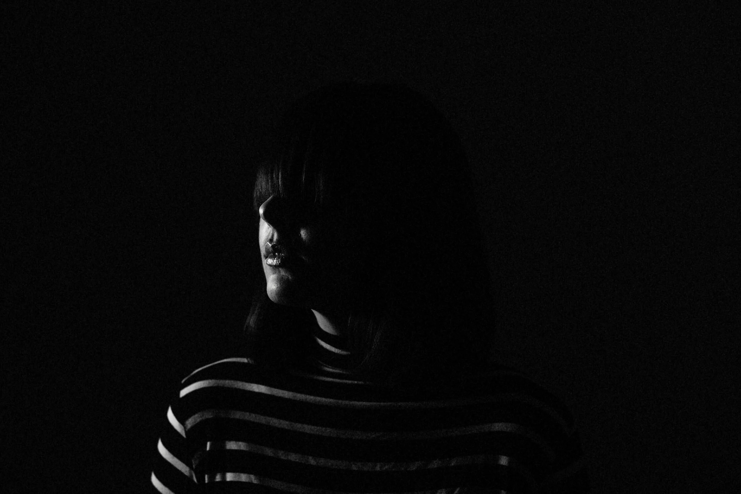 Side profile of a woman with dark hair wearing a striped shirt, illuminated from the side in a dark background.