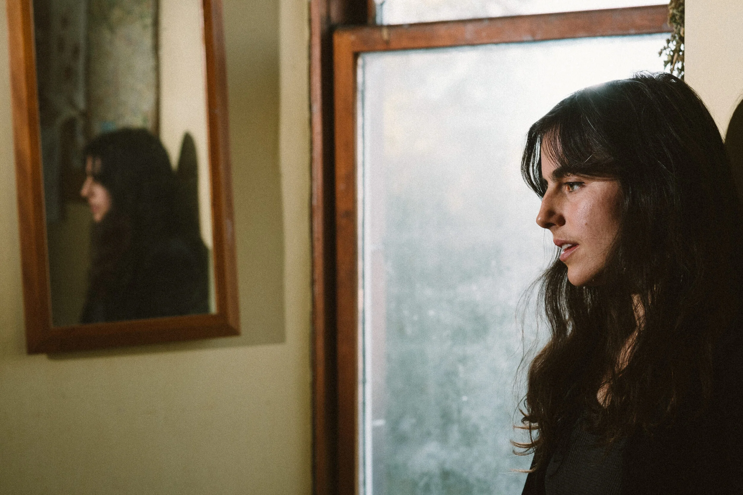 A woman with dark hair standing in front of a window with a wooden frame, looking to her right, with an image of herself reflected in a nearby mirror.