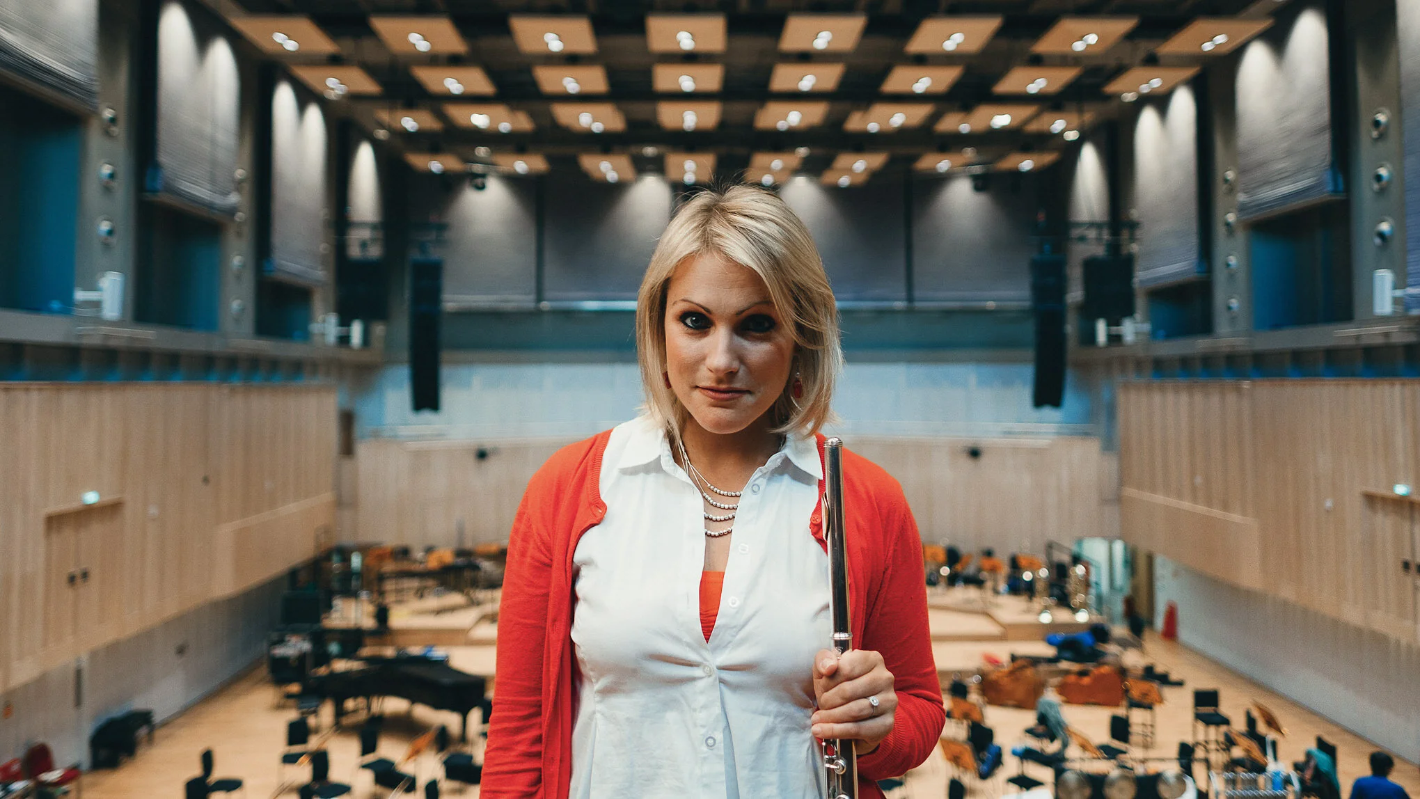 A woman standing in a large room with a high ceiling and wooden panels, holding a flute and looking directly at the camera.
