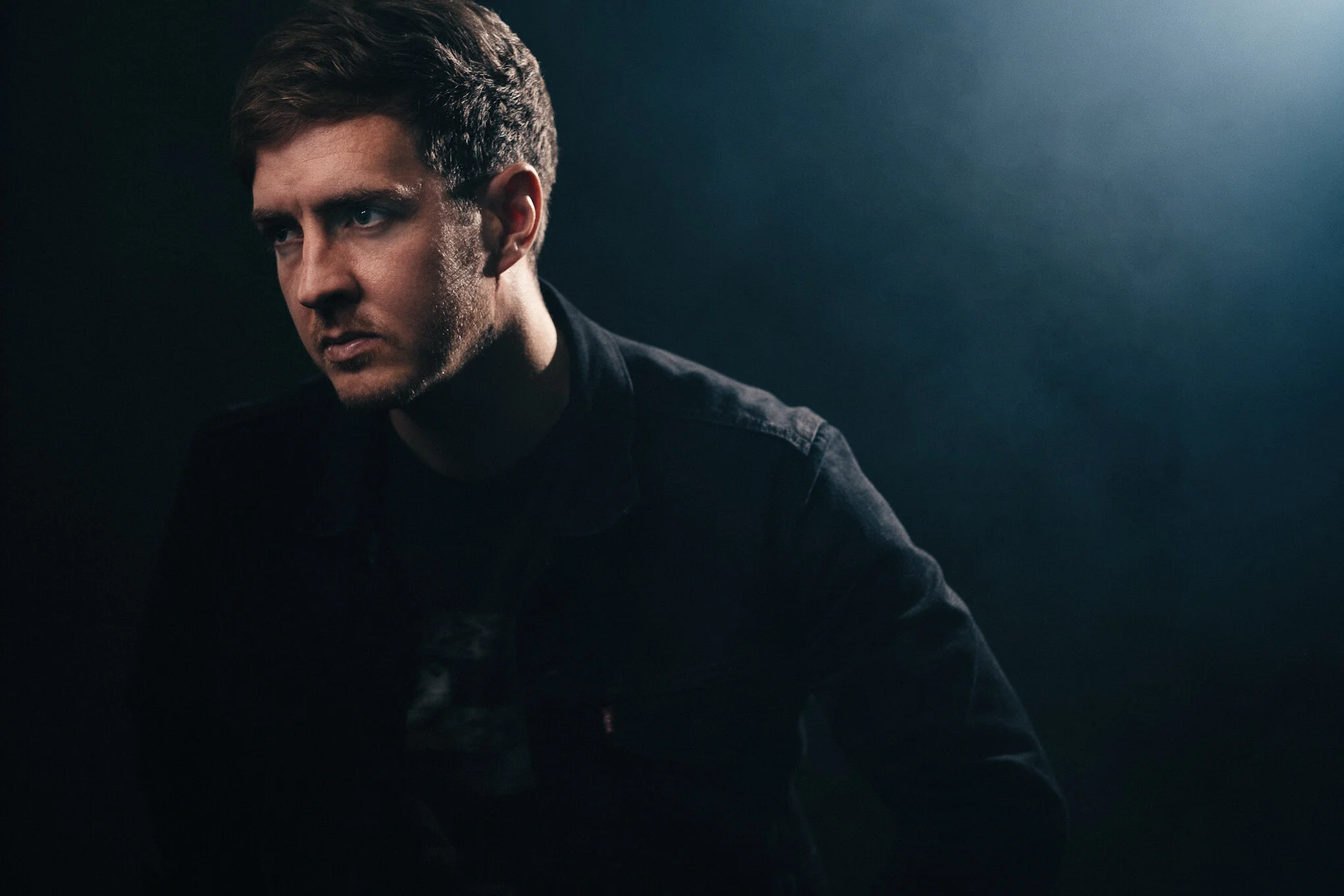 Close-up portrait of a young man with short dark hair, wearing a black shirt, looking to the side against a dark background with soft lighting.