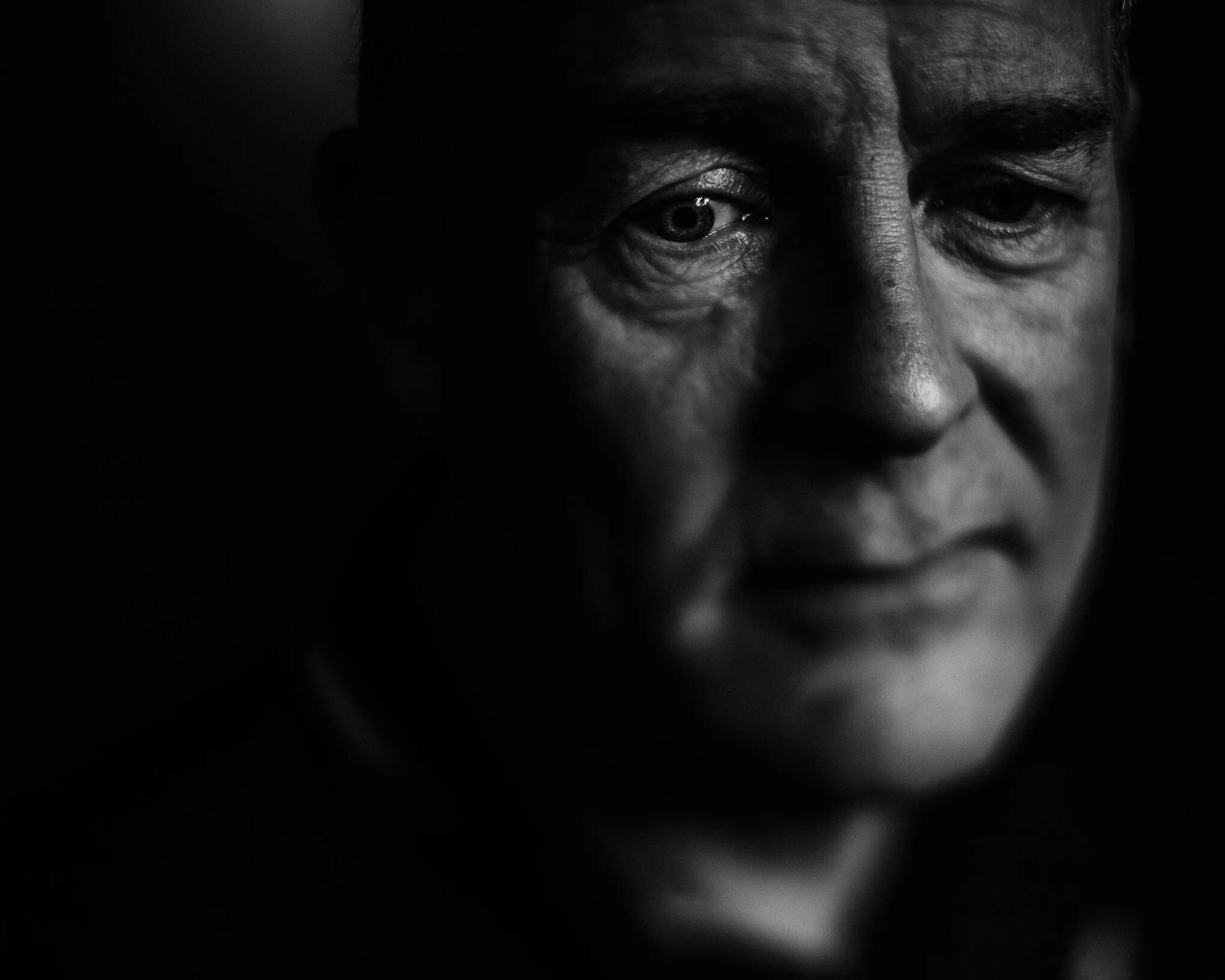 Close-up black and white photo of a man's face, focusing on his eyes and forehead, with deep shadows creating a dramatic effect.