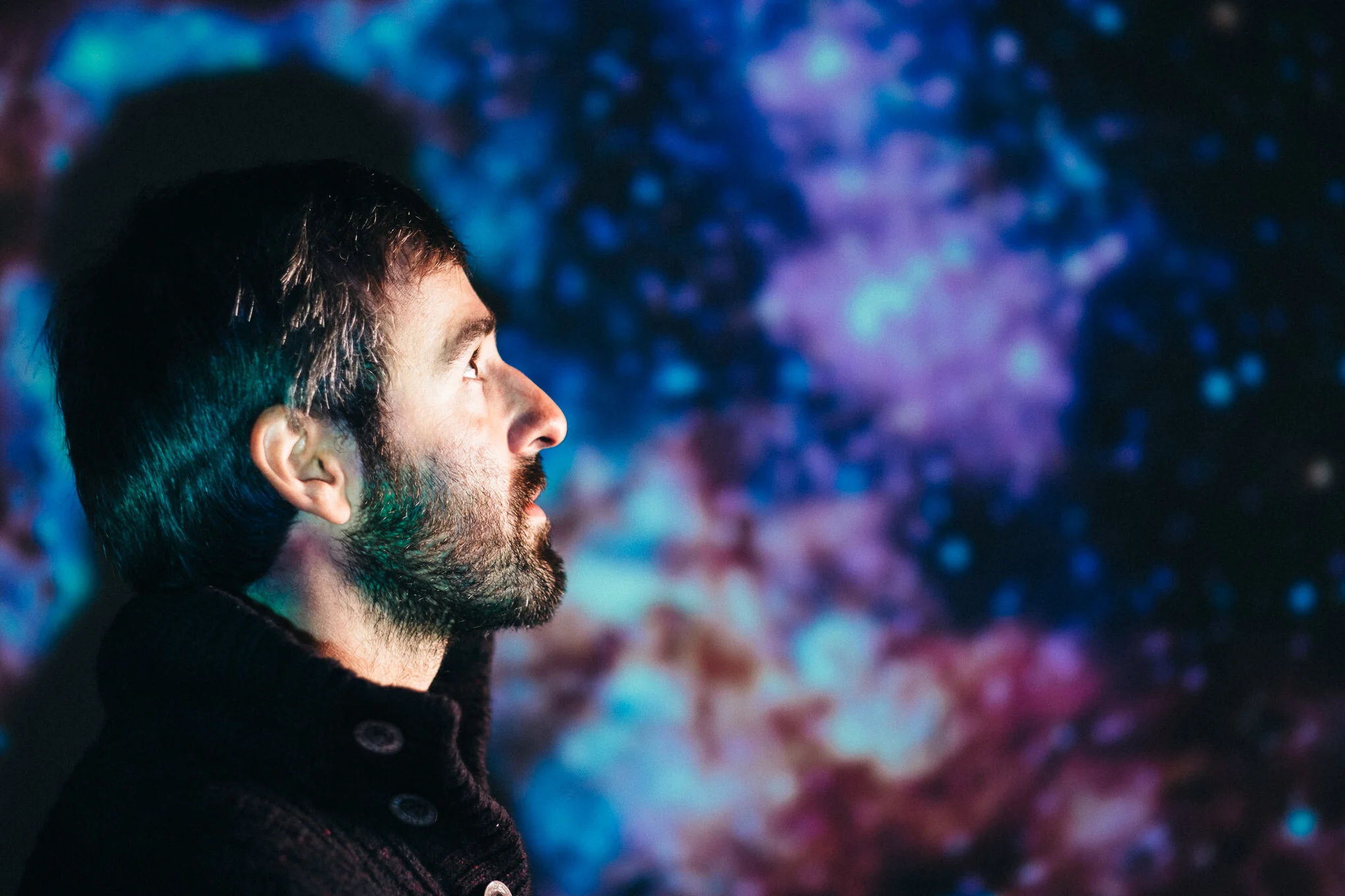 A man with dark hair and beard is looking at a colorful, abstract light display in the background.