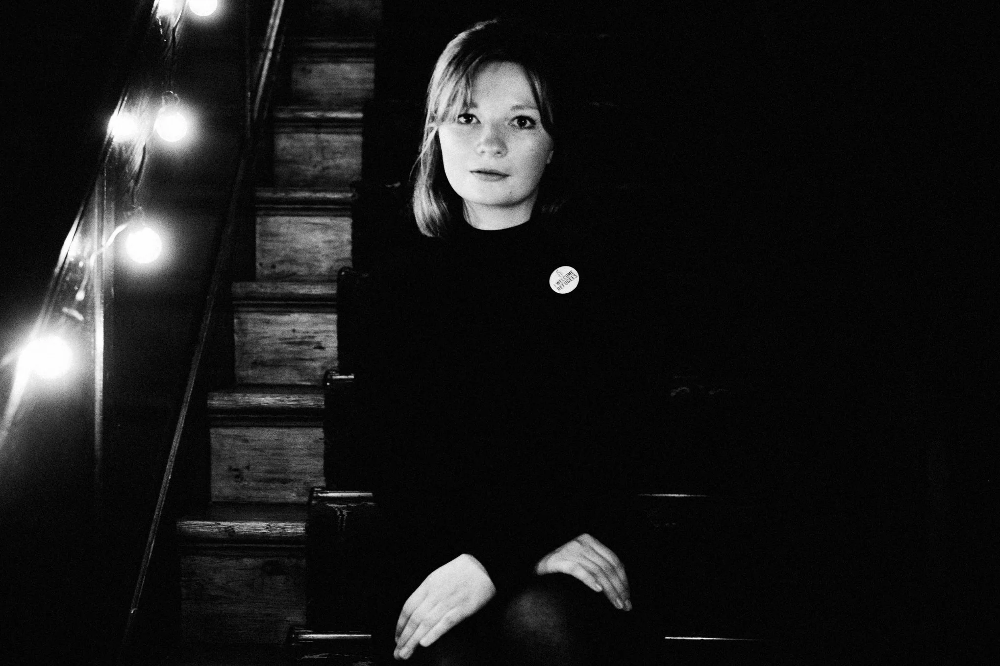 A black-and-white photo of a woman with shoulder-length hair, sitting at a piano with string lights reflecting off its surface. She is wearing a dark top and has a button that reads 'Hello Refugees' on her chest.