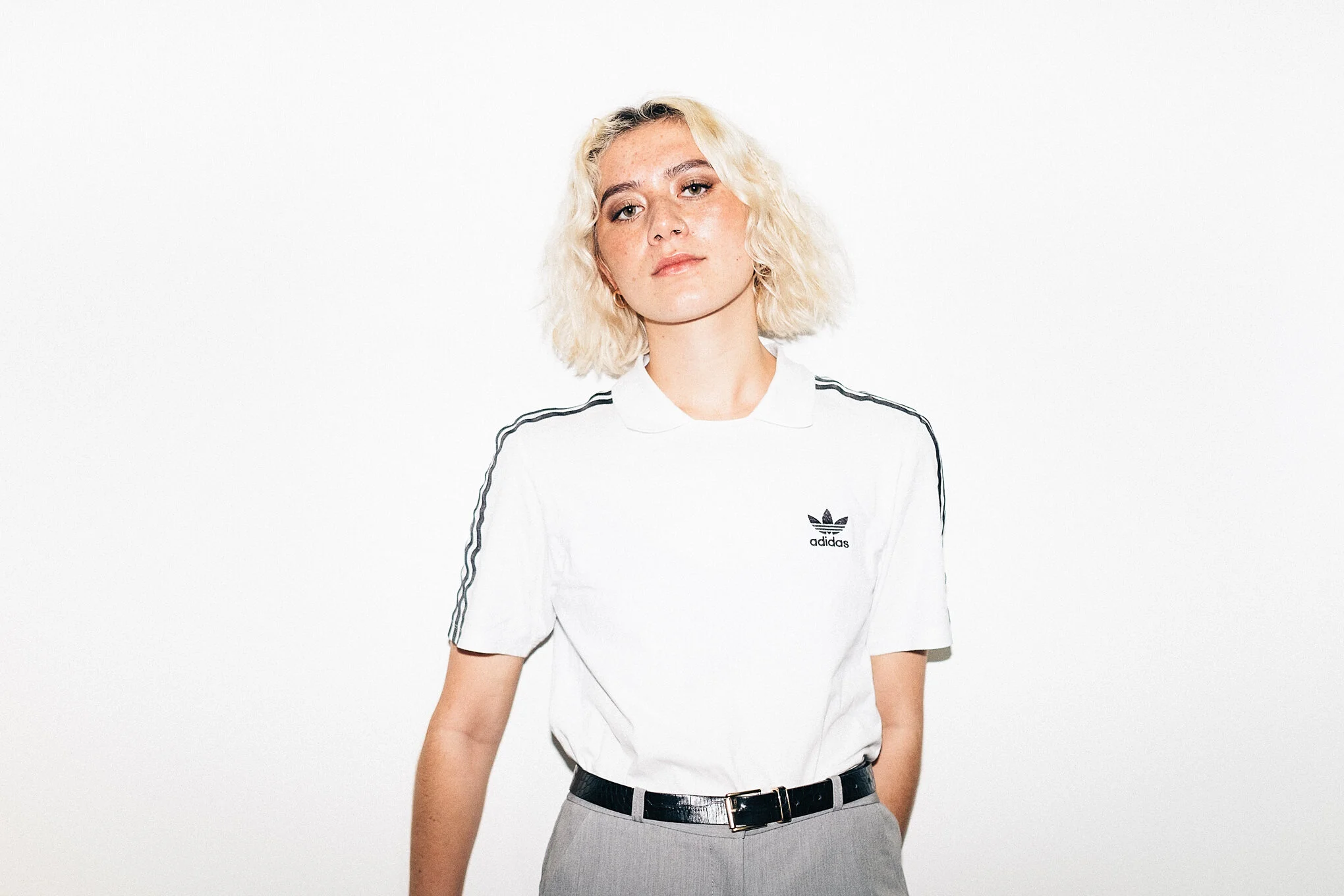 A young woman with platinum blonde hair wearing a white Adidas t-shirt with black stripes and gray pants standing against a white background.