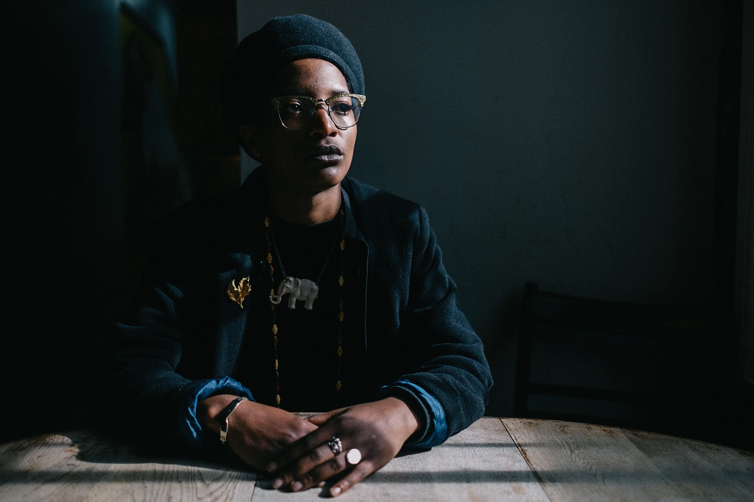 A woman with dark skin wearing glasses and a black head wrap sitting at a wooden table with her hands clasped, in a dimly lit room.