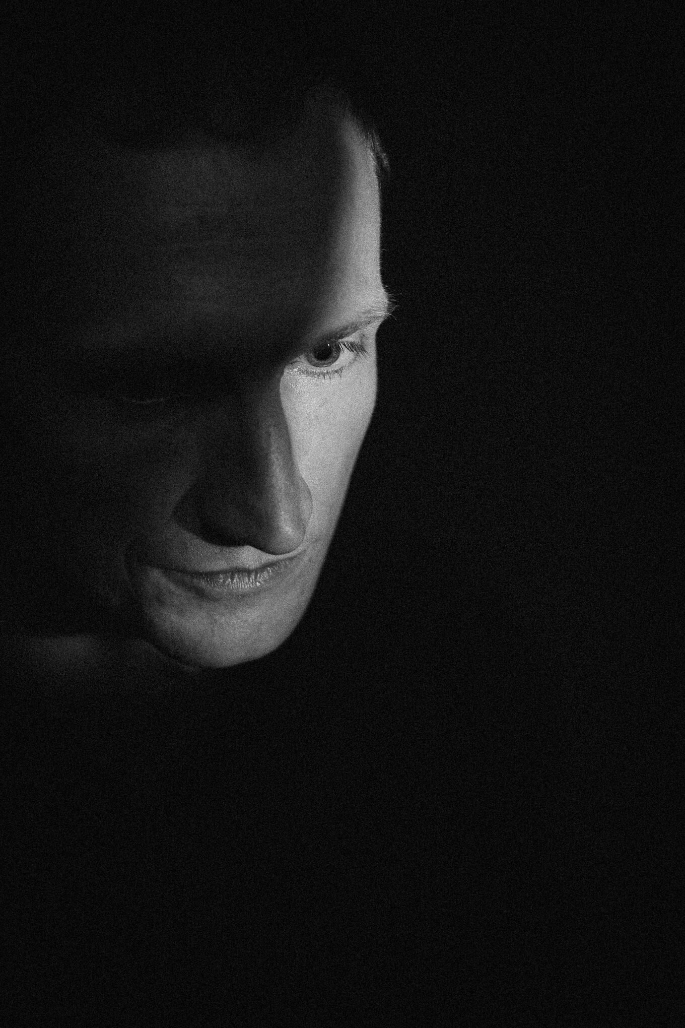 Close-up black and white photo of a person's face, highlighting the eye and part of the nose with shadows and soft lighting.
