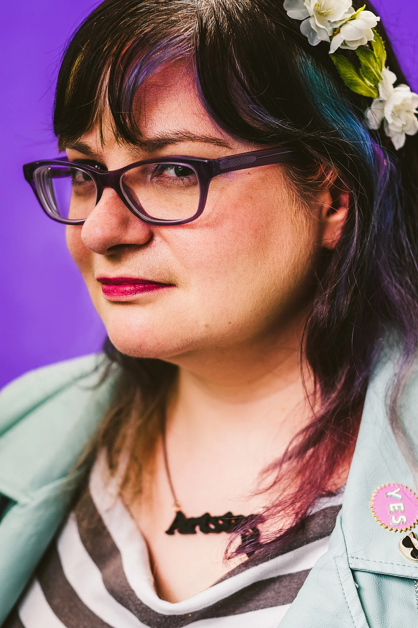 Close-up of a woman with dark hair, wearing glasses, earrings, and a flower headband, with a purple background.