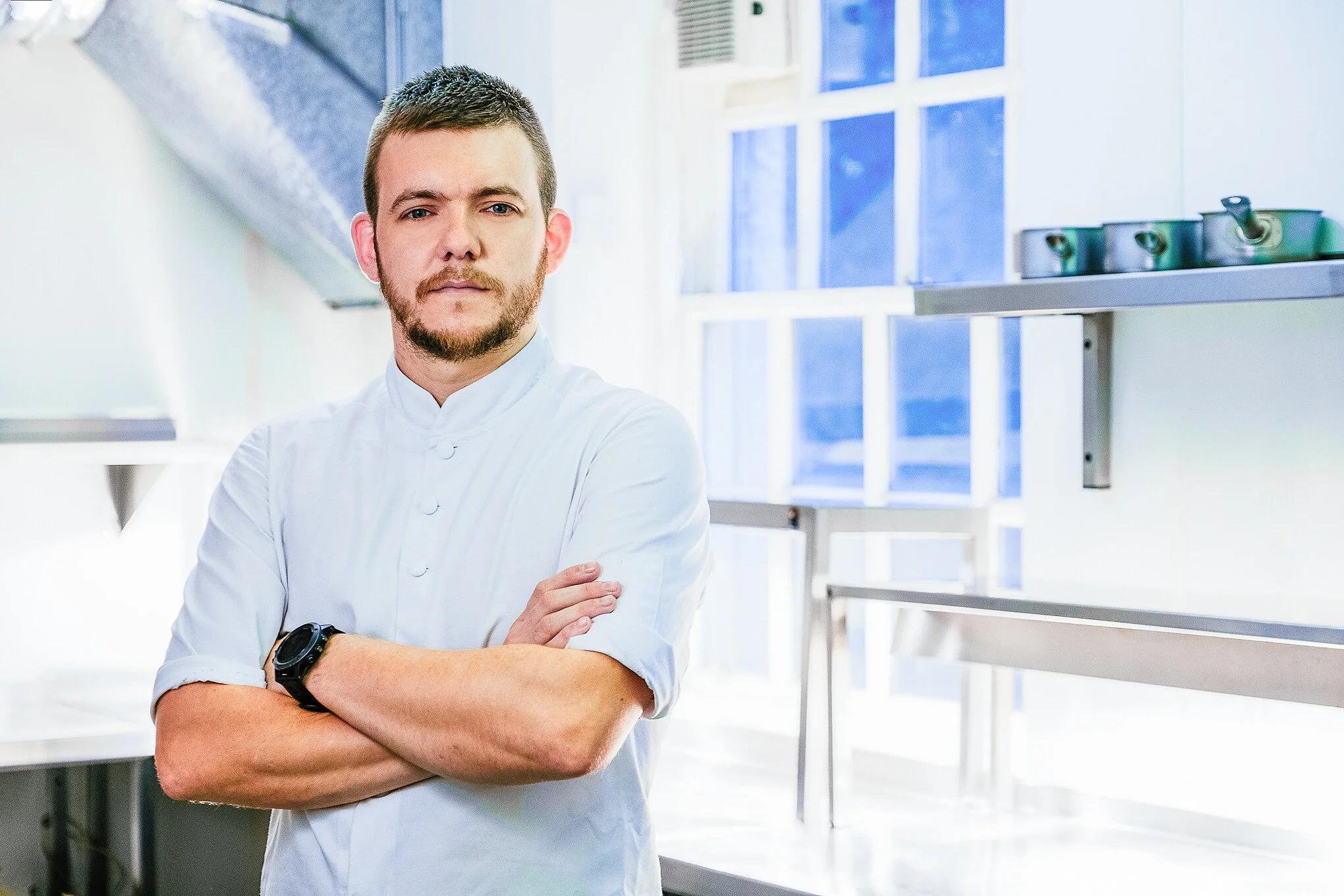 A young male chef with short hair, beard, wearing a white chef's coat, standing with arms crossed in a bright professional kitchen.