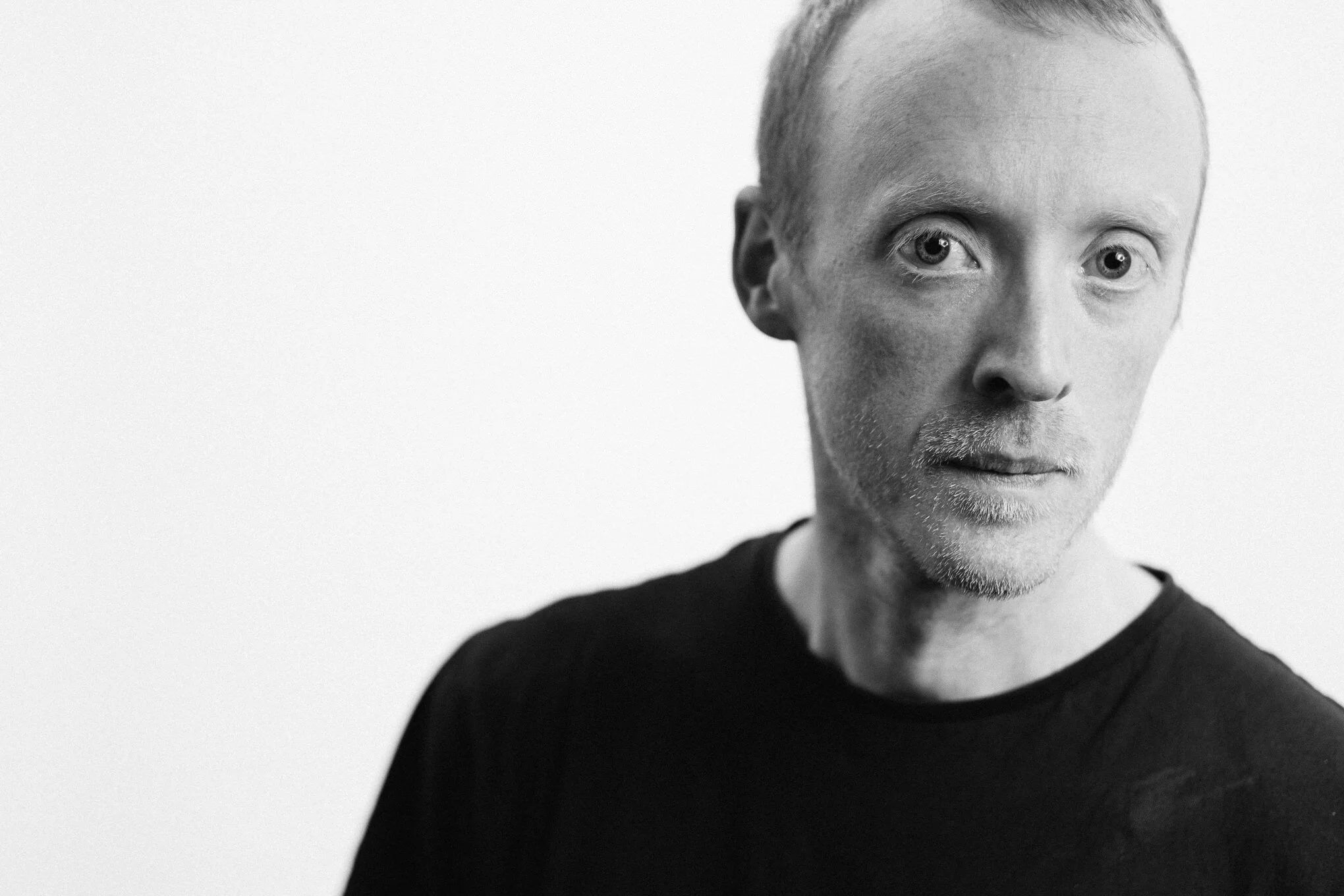 Close-up black and white portrait of a man with short hair, light beard, wearing a dark shirt, looking into the camera against a plain white background.