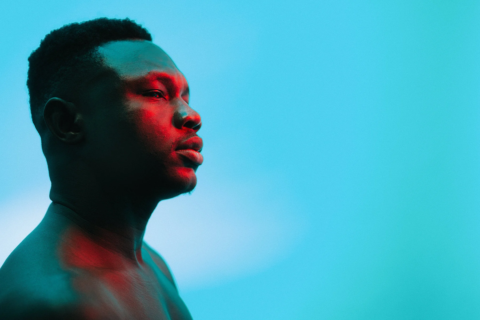 Profile of a young man with short hair looking to the right against a blue background, lit with red and blue lighting.
