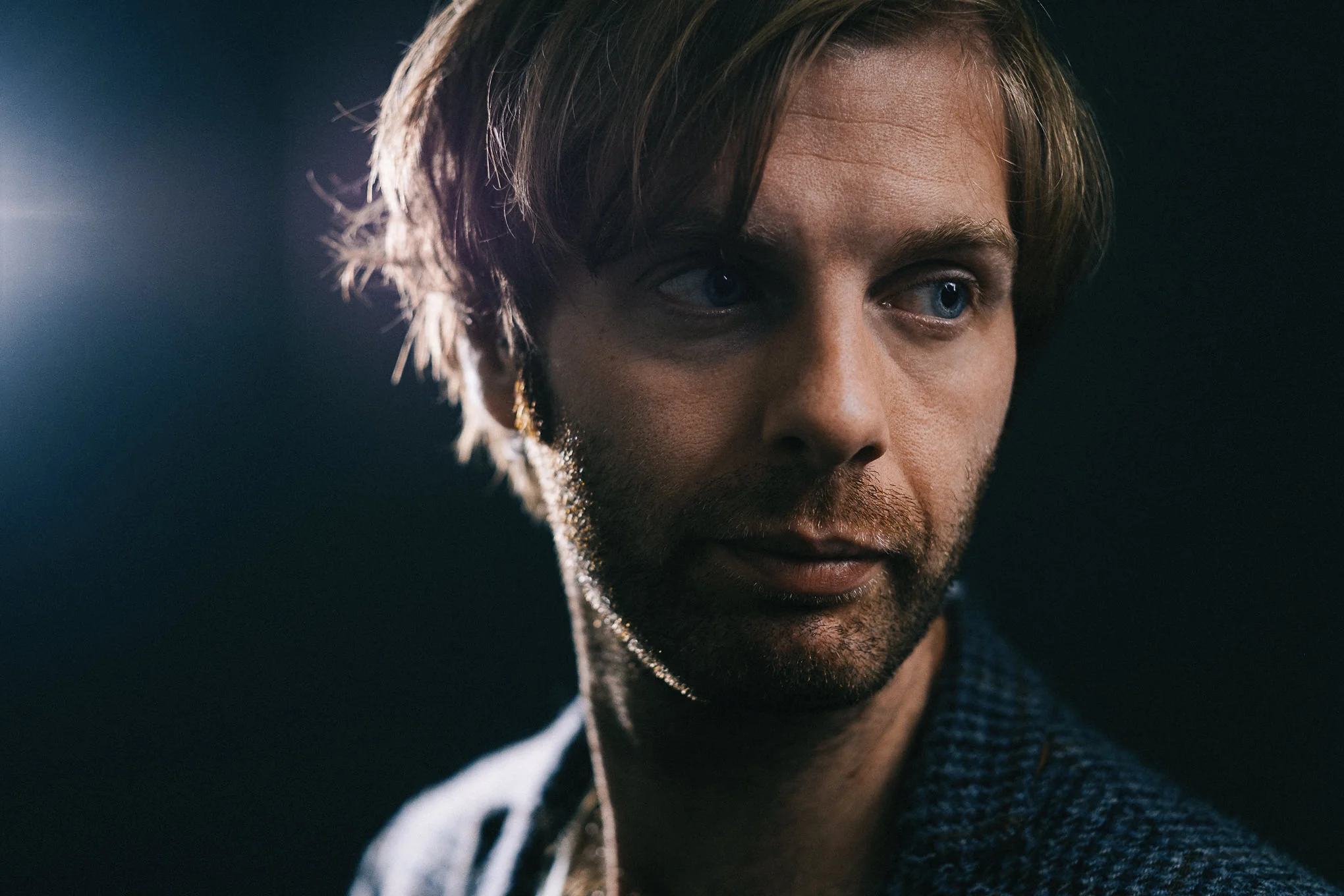 Close-up of a man's face with a serious expression, looking slightly downwards, with tousled brown hair, blue eyes, and a beard, wearing a dark checkered shirt, with soft lighting highlighting face details.