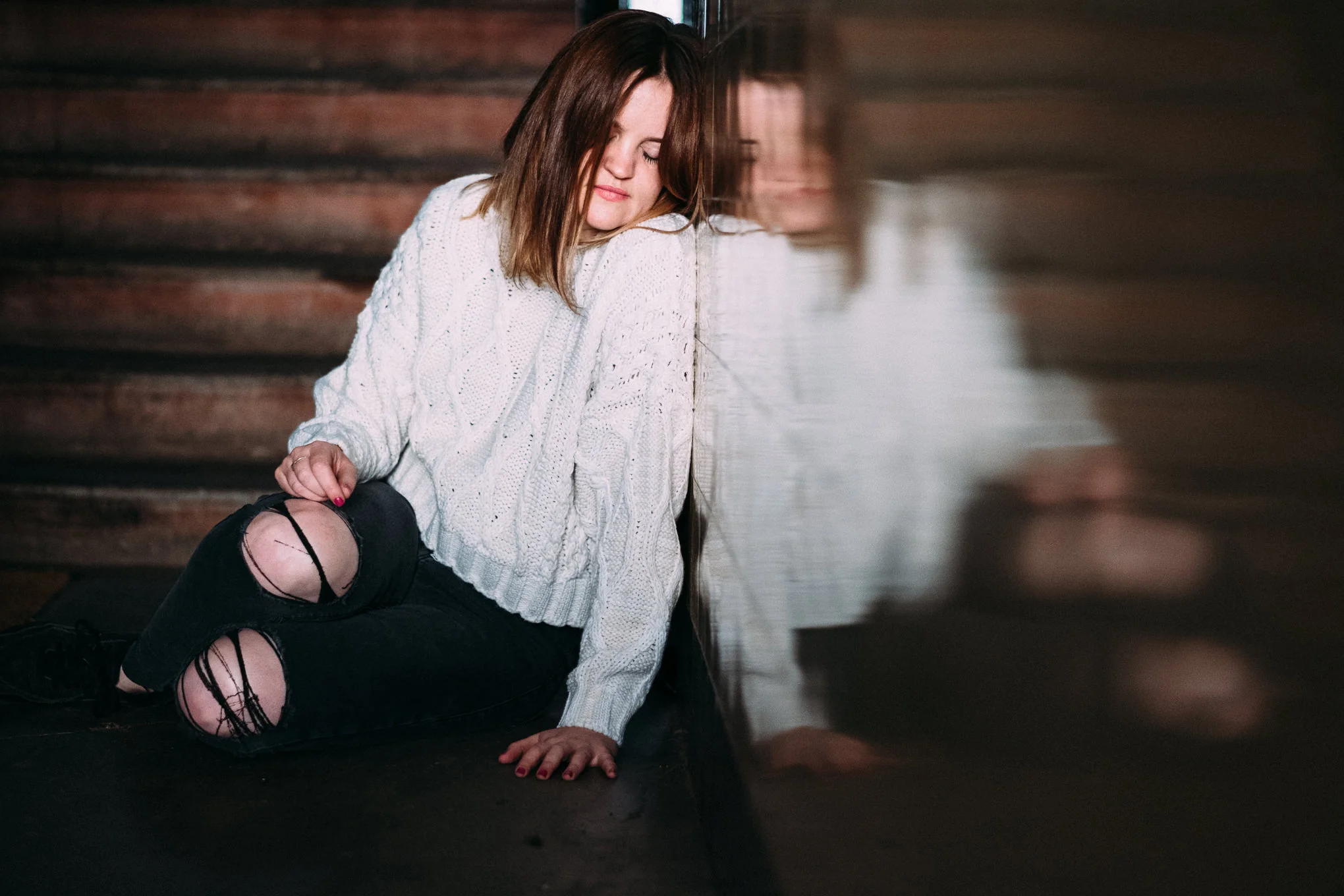 A young woman with brown hair and fair skin sitting on a dark floor leaning against a reflective wall, with her eyes closed. She is wearing a white cable-knit sweater and ripped black jeans. The setting appears to be indoors, with dark wooden stairs 