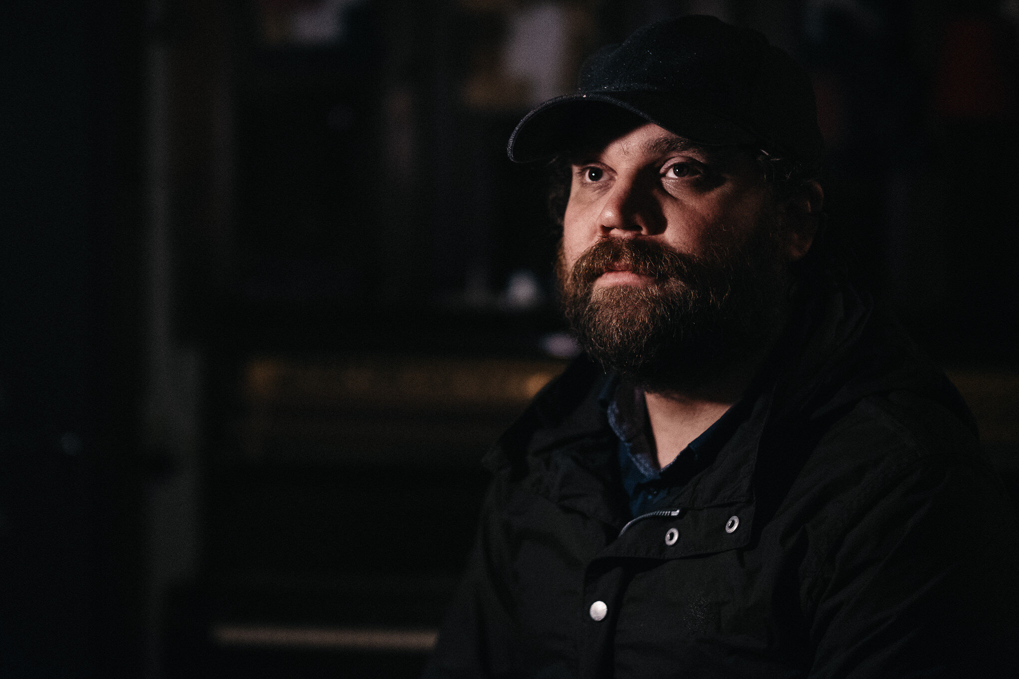 A man with a beard, wearing a black baseball cap and black jacket, looking thoughtfully into the distance in a dimly lit environment.